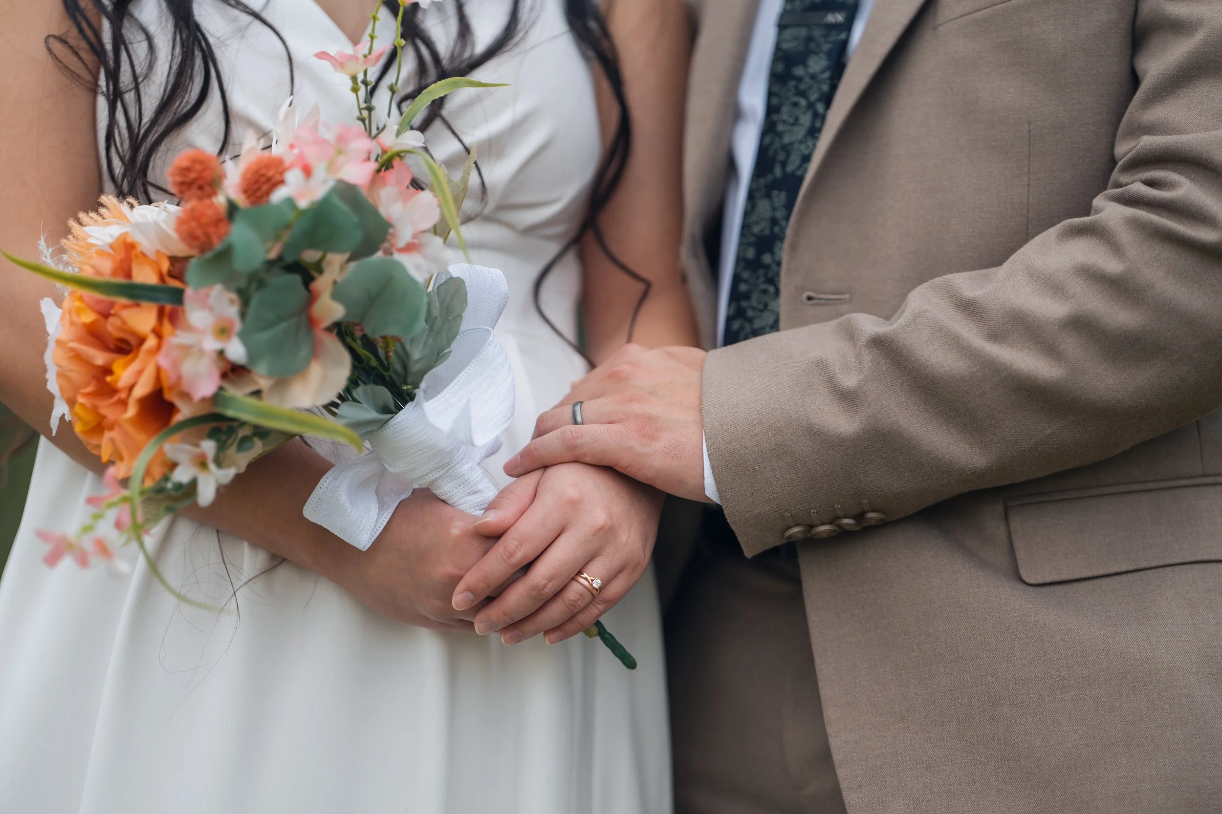 Close-up of a bride and groom holding hands during a wedding ceremony, with the bride holding a bouquet of flowers and both wearing wedding rings.