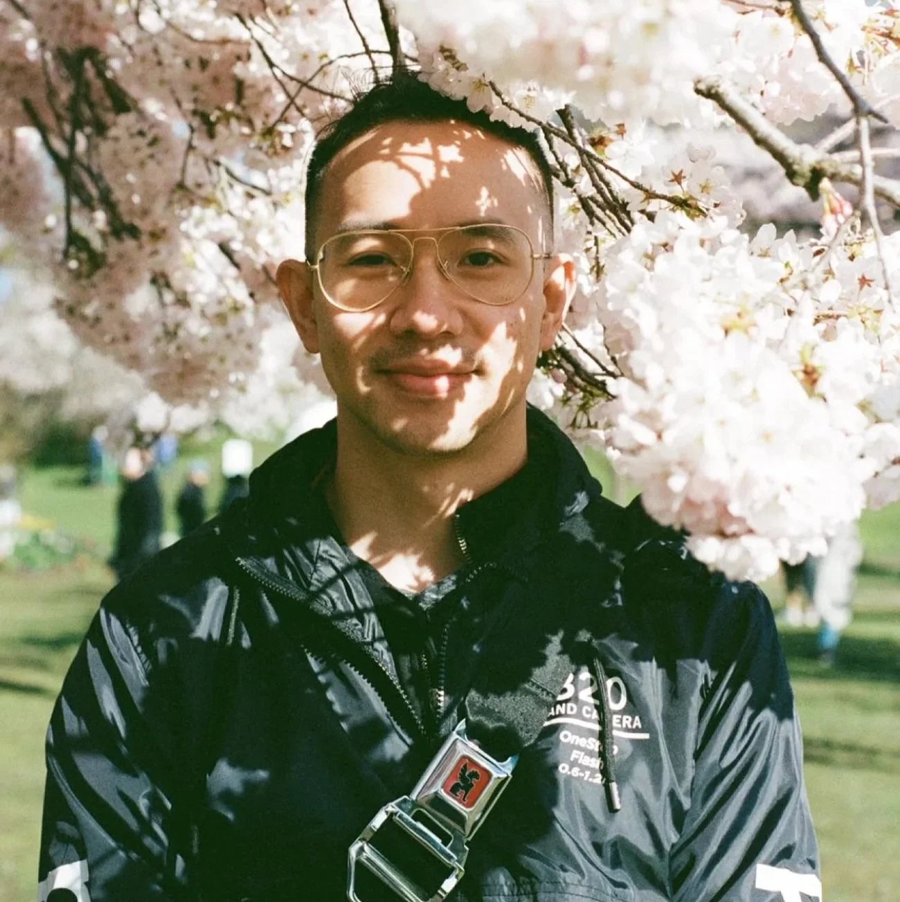 A young man smiling, standing outdoors near cherry blossoms, wearing glasses and a black jacket with a camera strap around his neck.