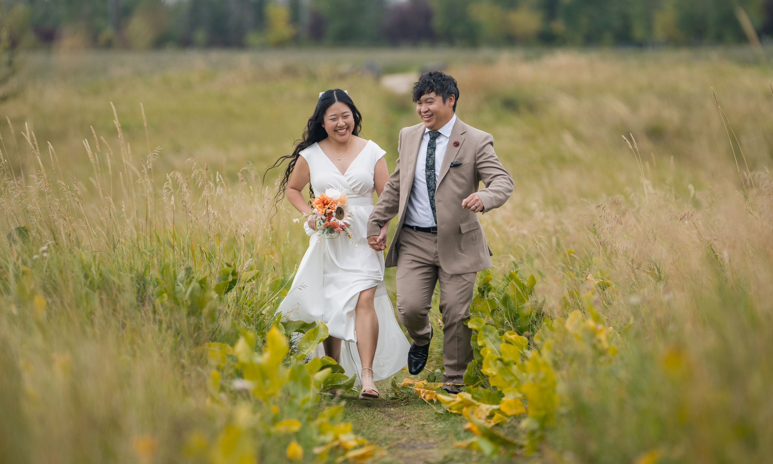 A smiling couple, a bride in a white dress holding a bouquet and a groom in a beige suit, running hand-in-hand through a grassy field with trees in the background during daytime.