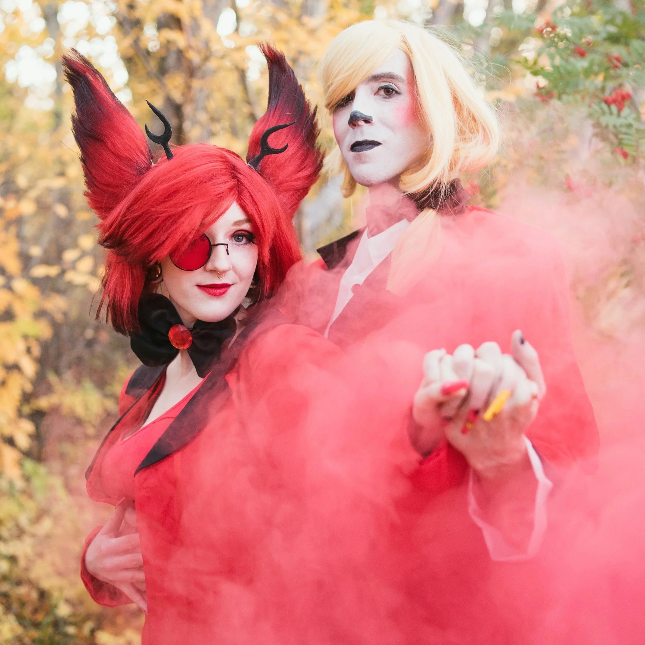 Two people dressed in Halloween costumes, one with fox-like features and red hair, the other with pale face makeup and blonde hair, surrounded by autumn trees and a pink smoke or fog.