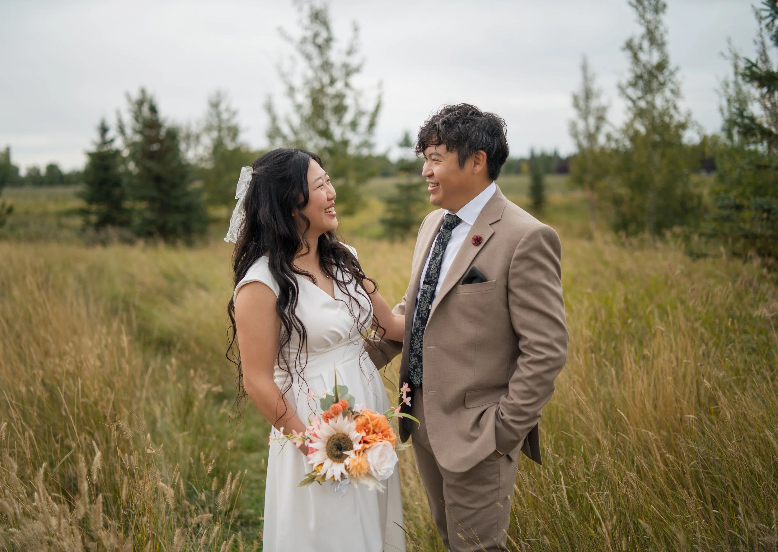 A bride and groom standing in a grassy field, smiling at each other, with trees and a cloudy sky in the background. The bride is holding a bouquet of flowers.
