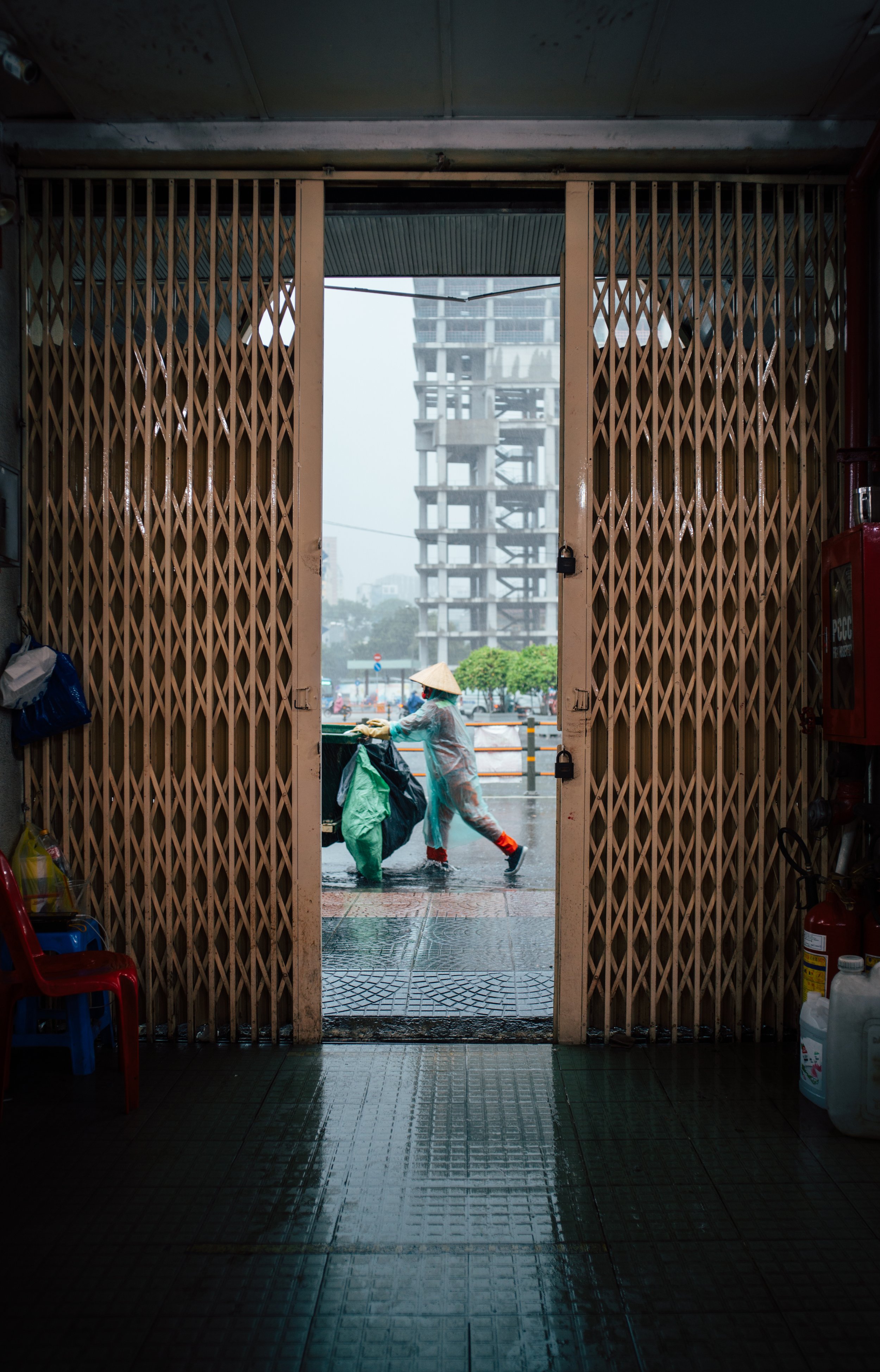 Person wearing raincoat and conical hat pushing a trash bin on wet street during rainy day