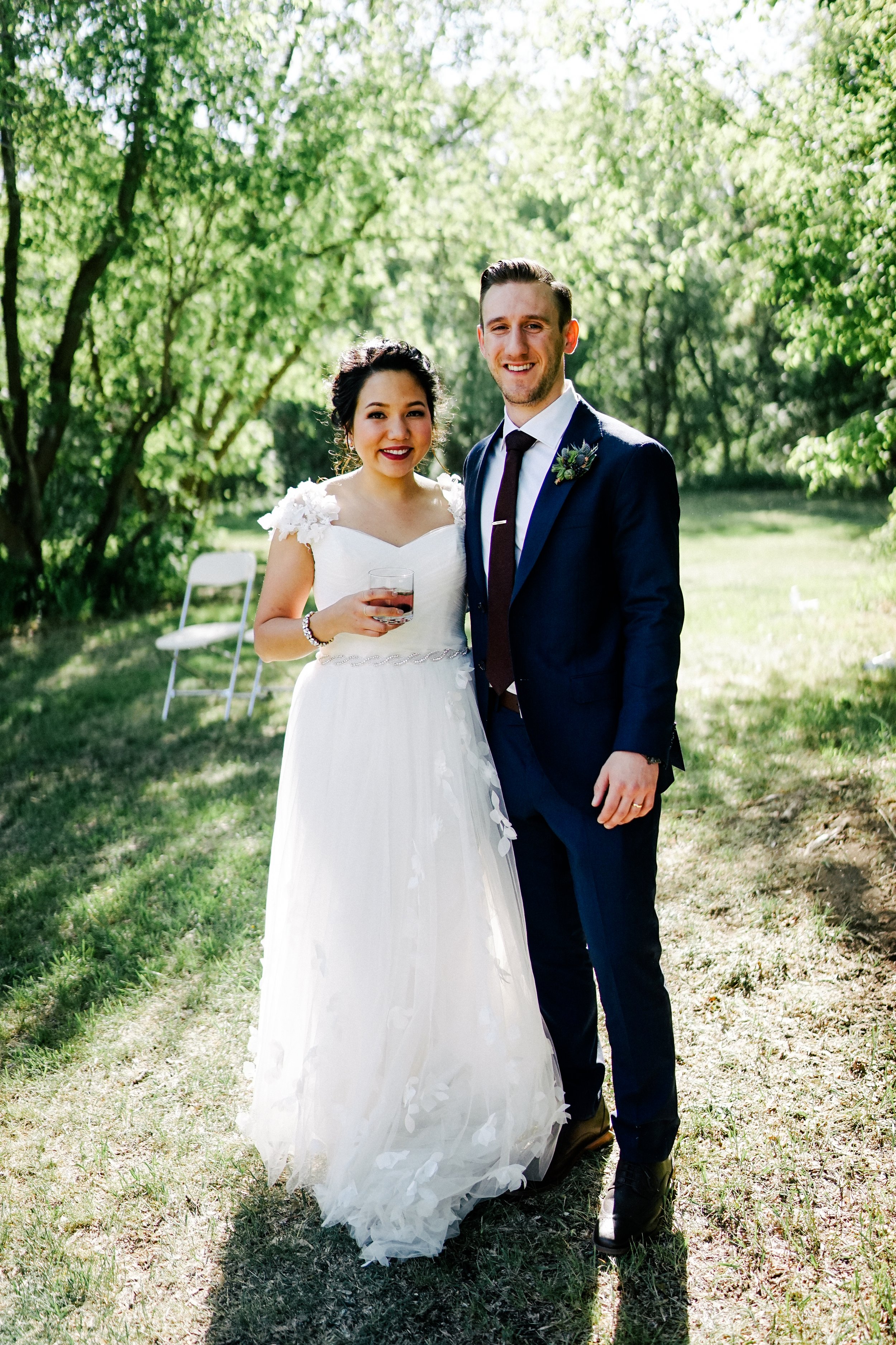 A bride and groom smiling outdoors on their wedding day, with trees and green foliage in the background. The bride is wearing a white wedding dress with floral details and holding a glass, while the groom is in a dark suit with a tie and boutonniere.