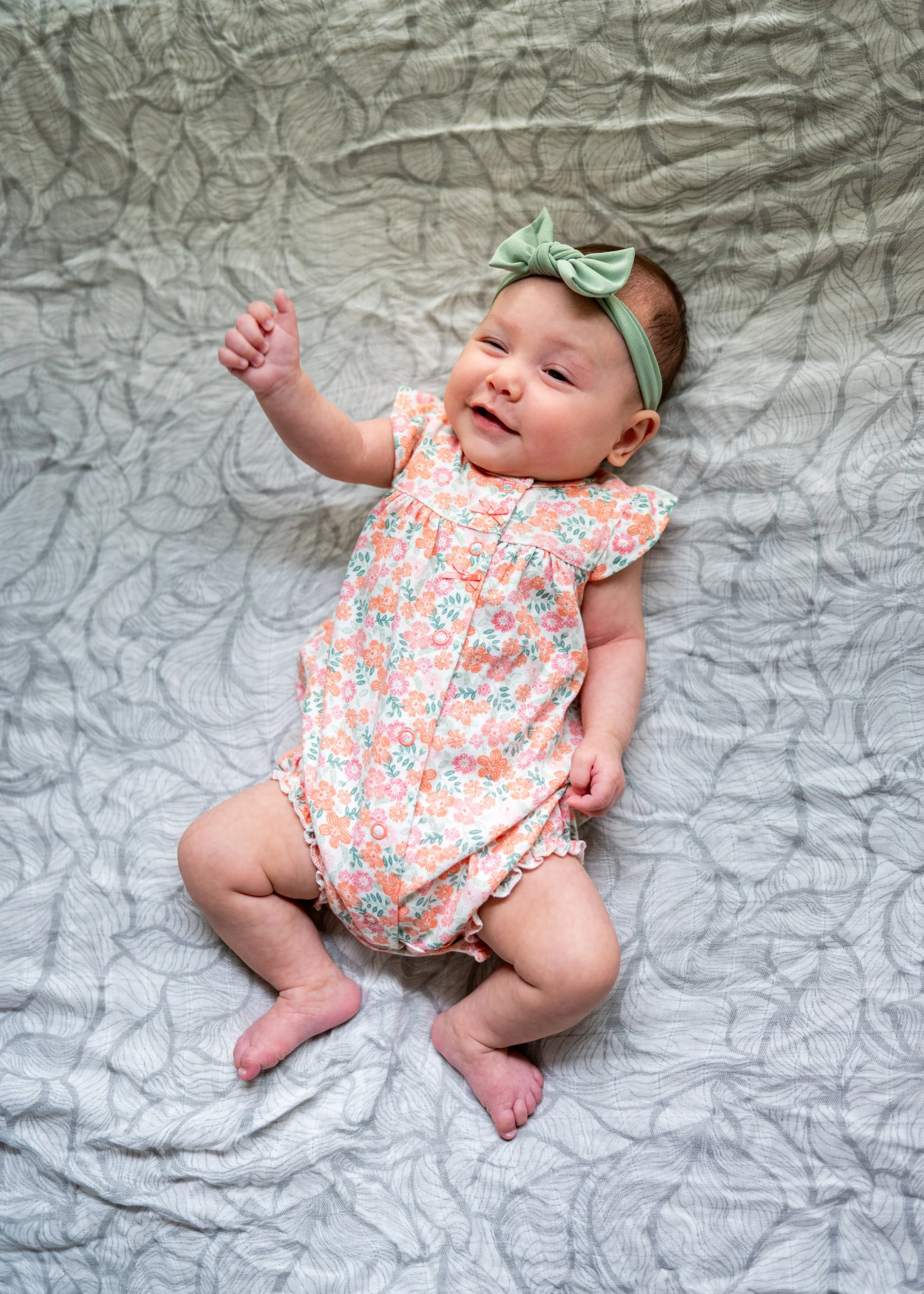 A baby girl lying on a light grey bedspread, wearing a floral dress and a green headband with a bow, smiling and raising one fist.