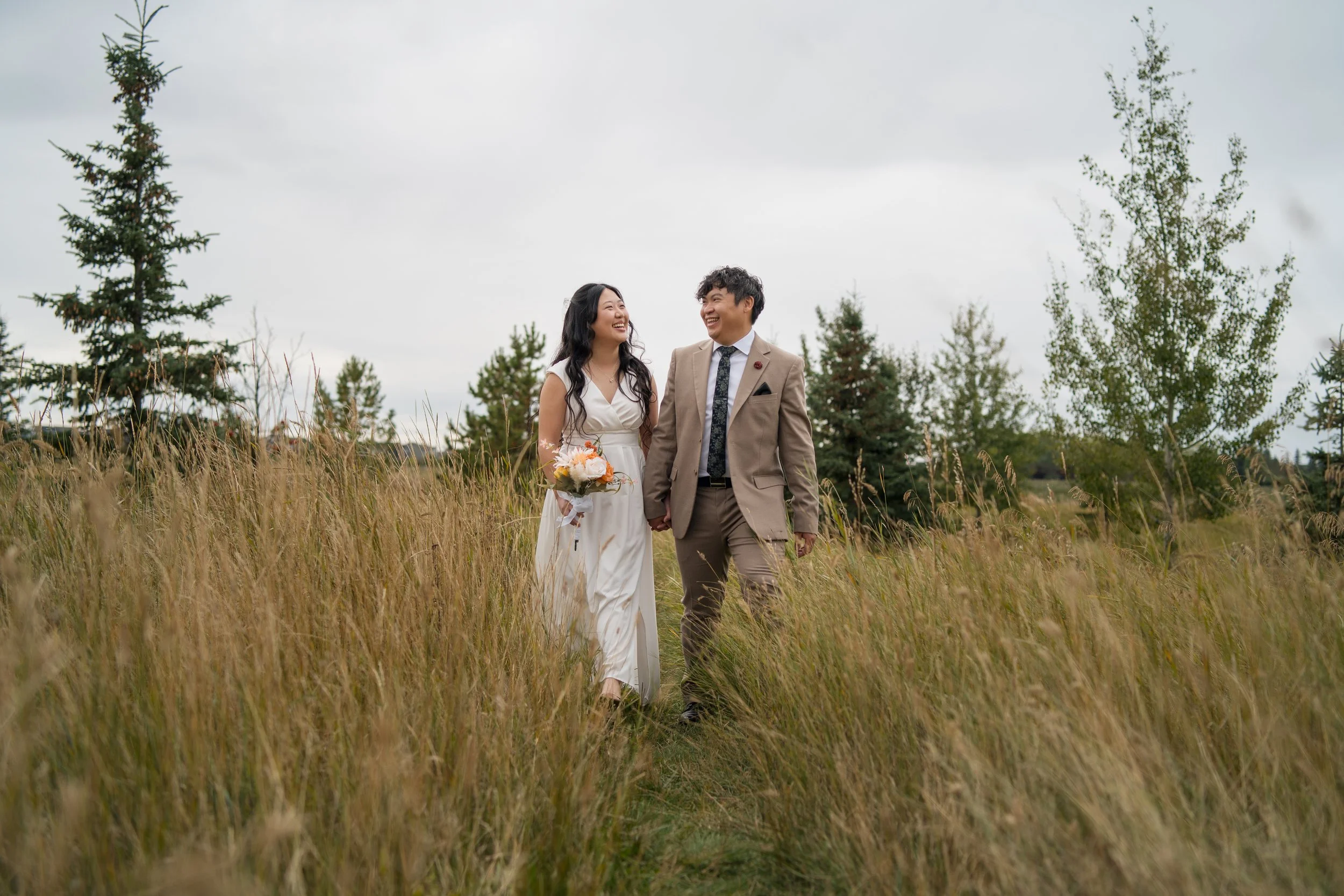 A newlywed couple walking hand in hand through a grassy field on a cloudy day, smiling and looking at each other, with trees in the background.