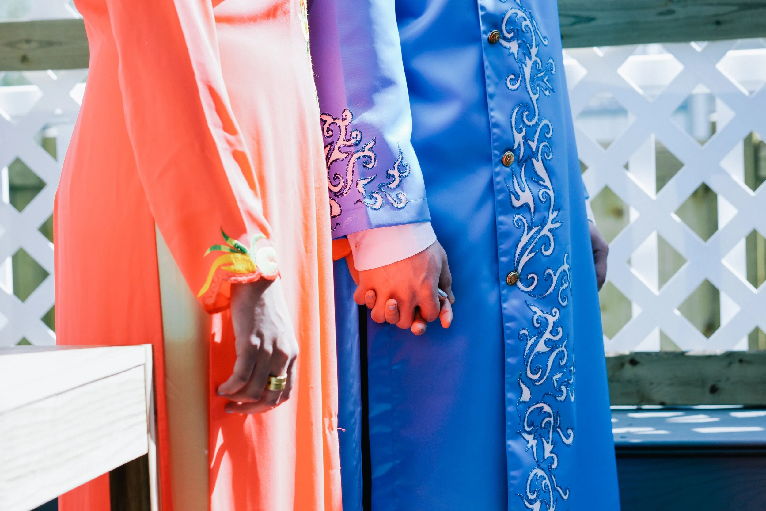 Close-up of two individuals holding hands, one wearing an orange dress with embroidered cuffs and the other wearing a blue garment with decorative embroidery, standing outdoors on a deck with a white lattice fence in the background.
