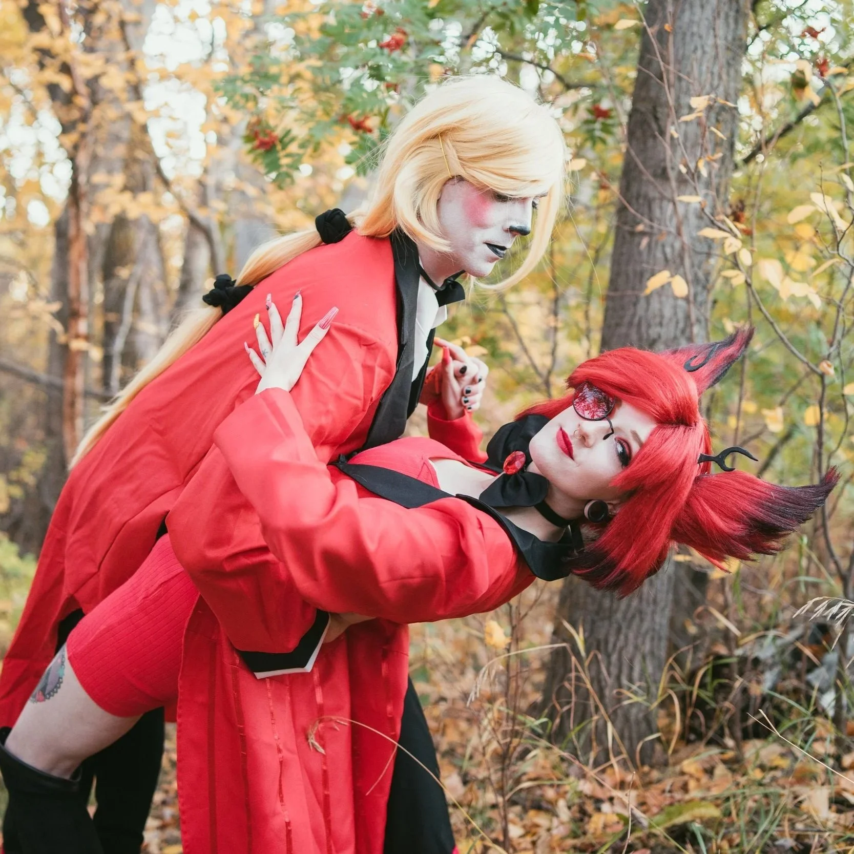 Two people in elaborate costumes with fantasy makeup posing outdoors in a wooded area with fallen leaves.
