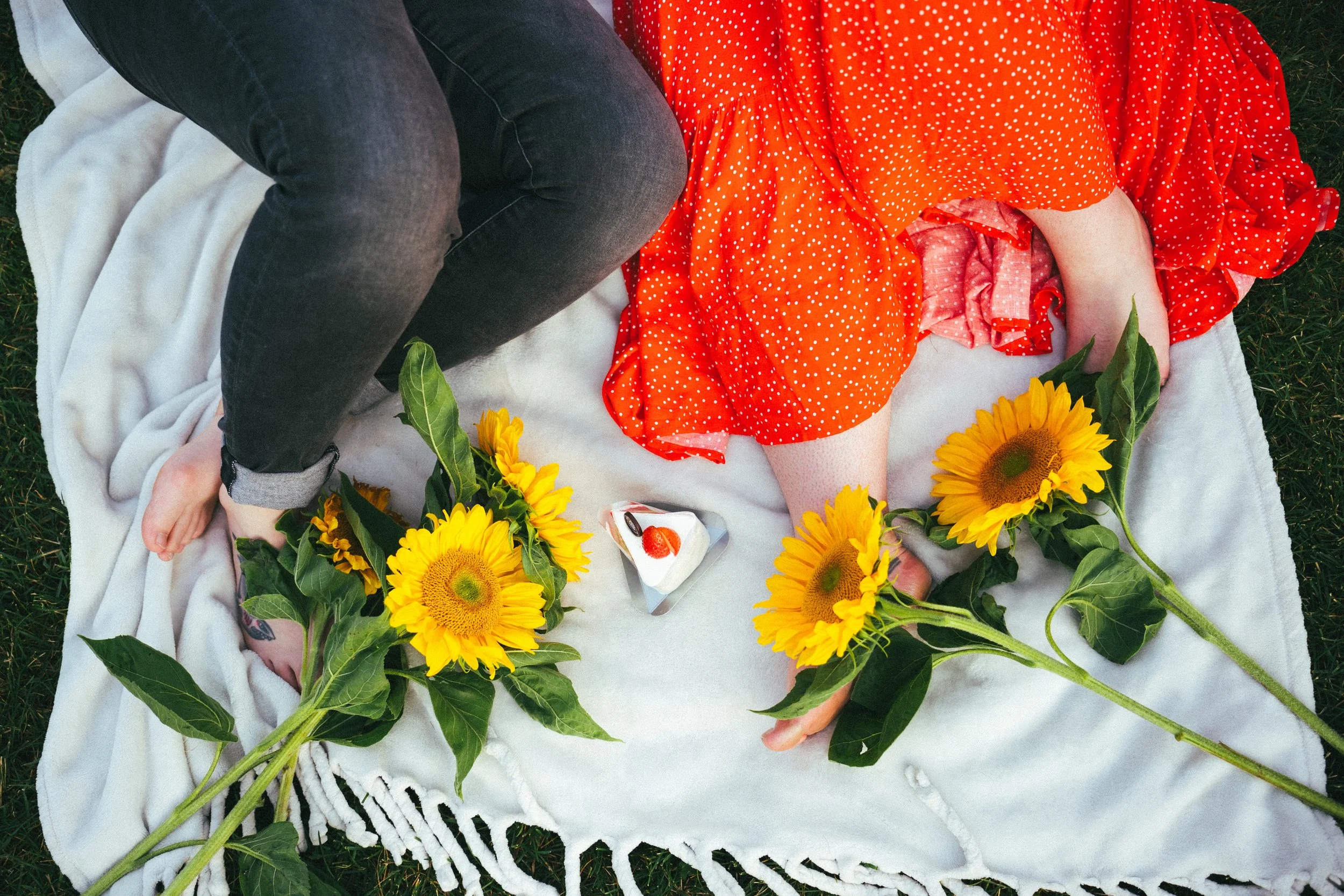A top-down view of two people lying on a white blanket outdoors. One person is in black jeans and the other in an orange dress. There are four large sunflowers placed on the blanket and a small piece of cake with strawberries.