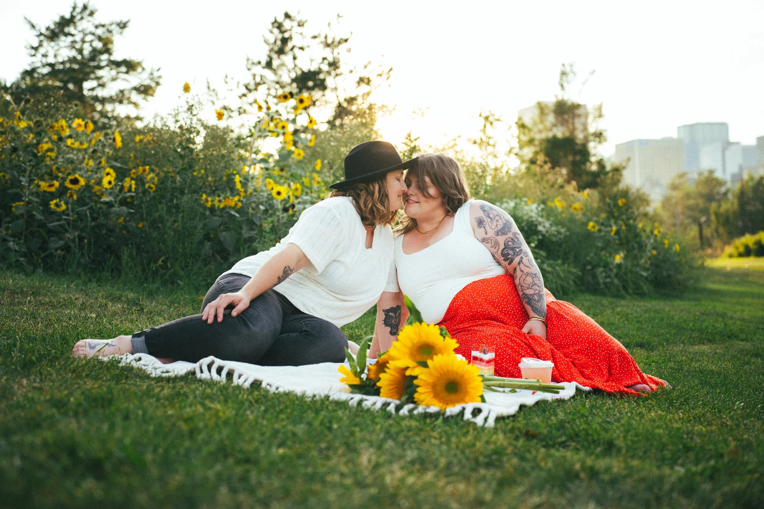 Two women sitting on a blanket in a park with sunflowers, leaning close and touching noses, with tall trees and city buildings in the background, during sunset.