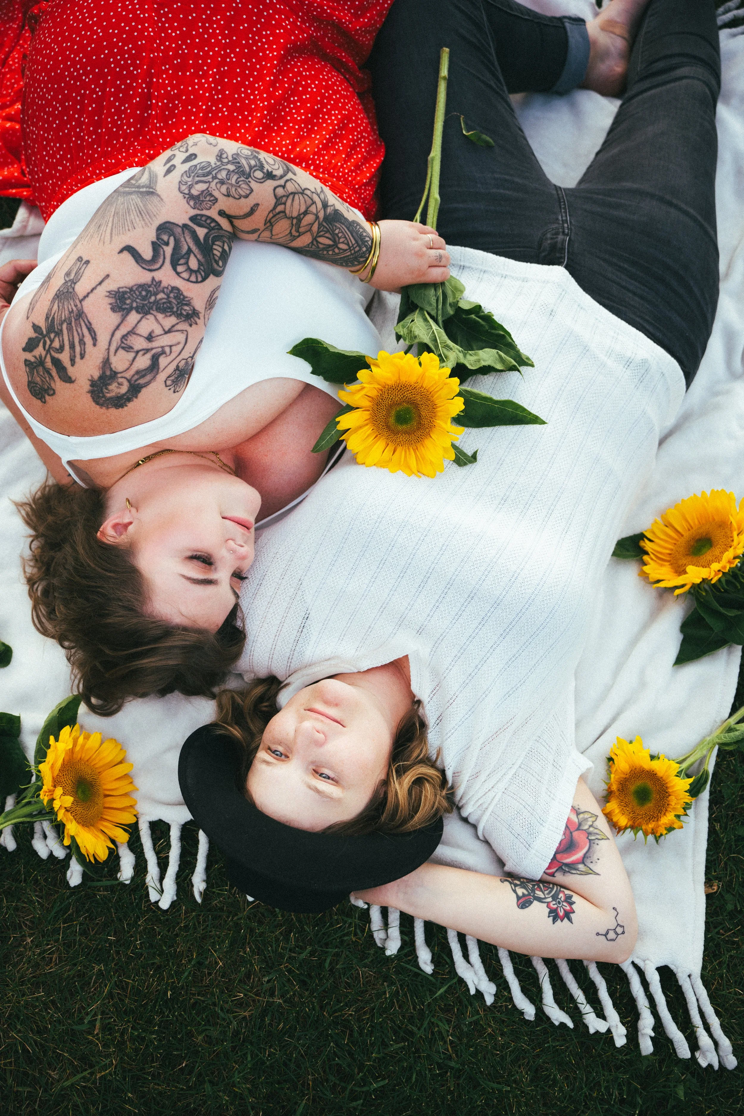 Two women lying on a blanket outdoors, surrounded by sunflowers. One woman is wearing a white top, a black hat, and has tattoos on her arms, while the other is wearing a red shirt with white polka dots, also with tattoos. They are relaxing and looking at each other.