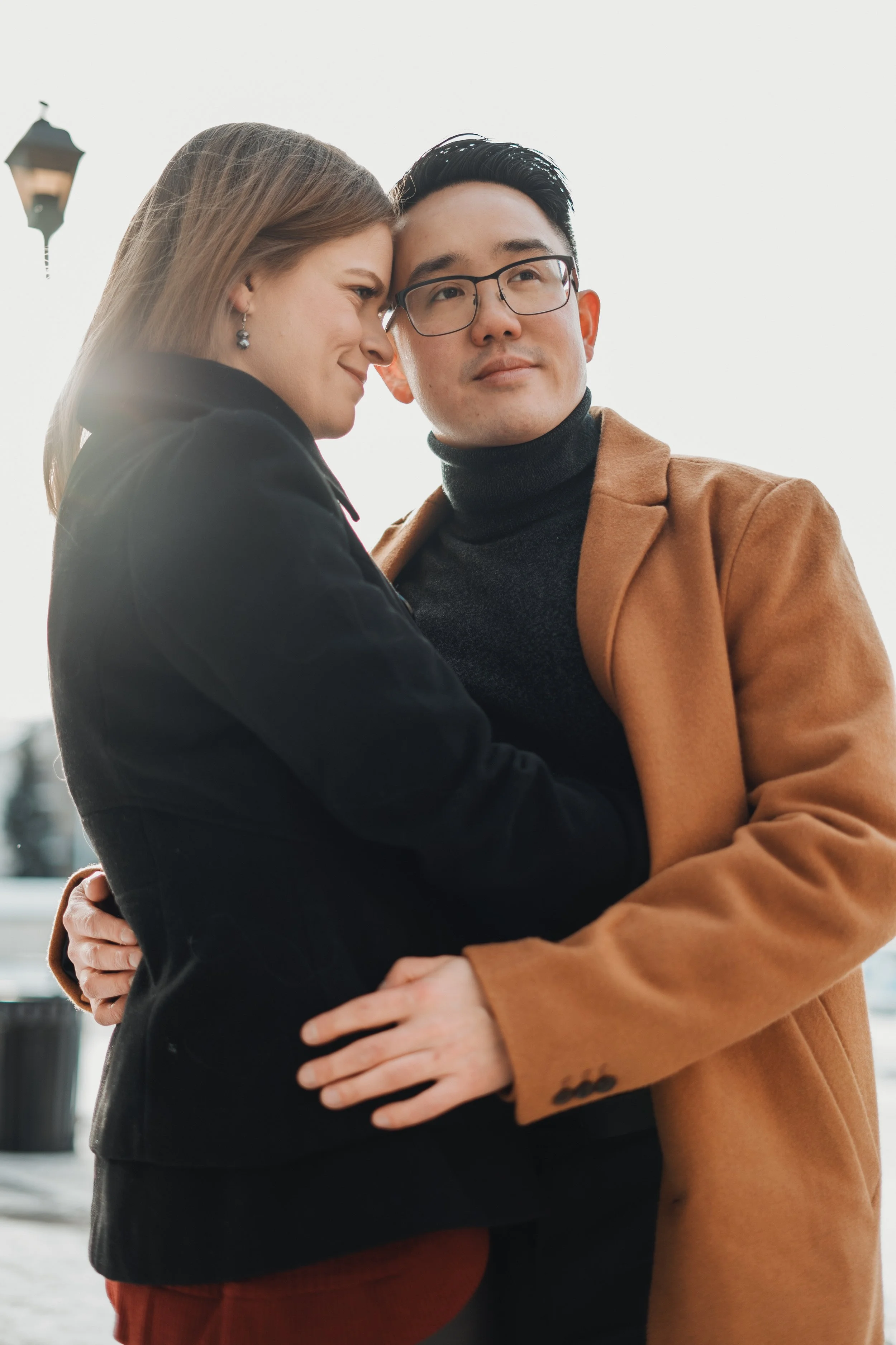 A couple embracing outdoors, with the woman smiling and the man looking off into the distance, during daytime.