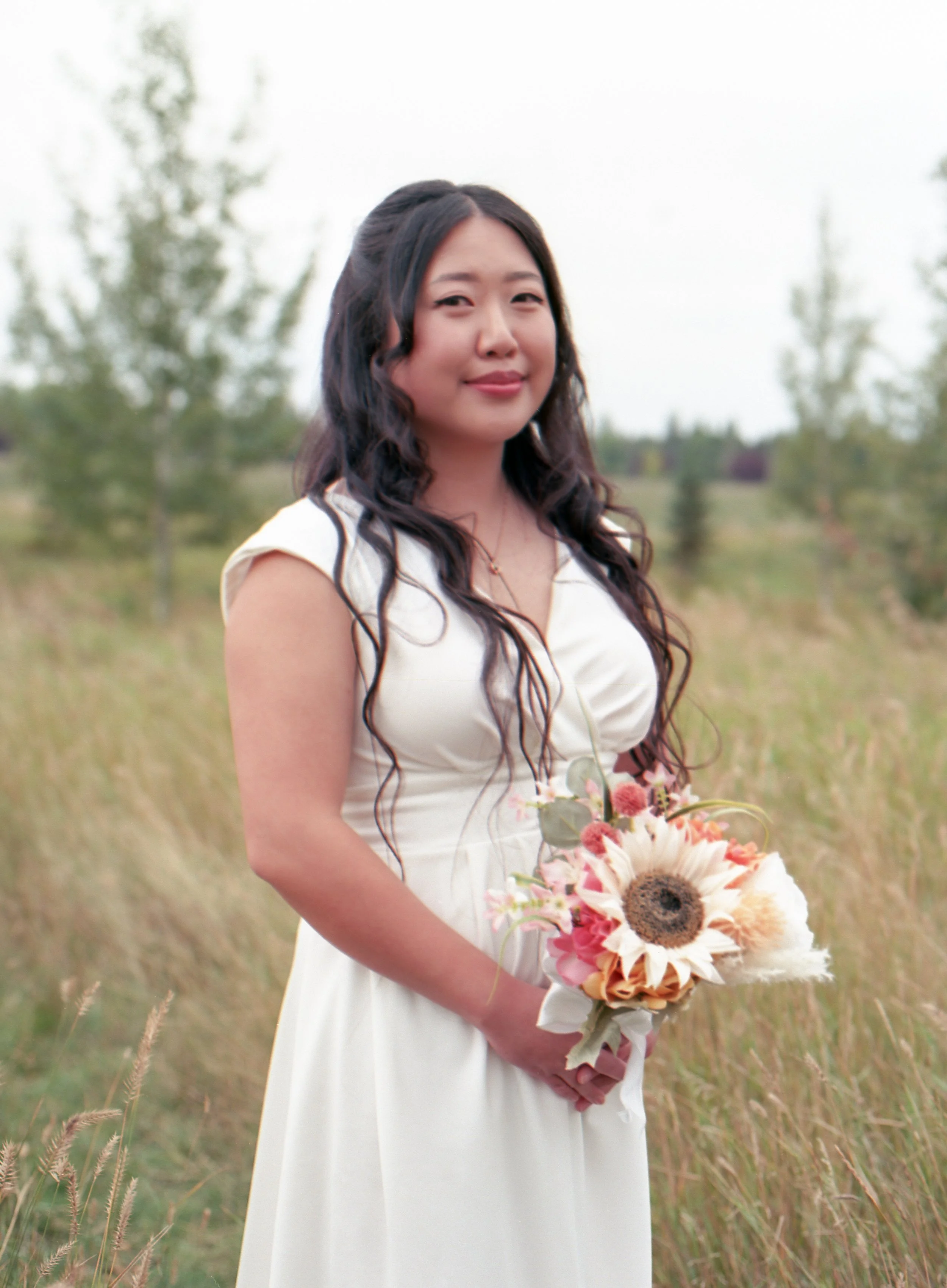 A woman in a white dress holding a bouquet of flowers outdoors in a grassy field with trees in the background.