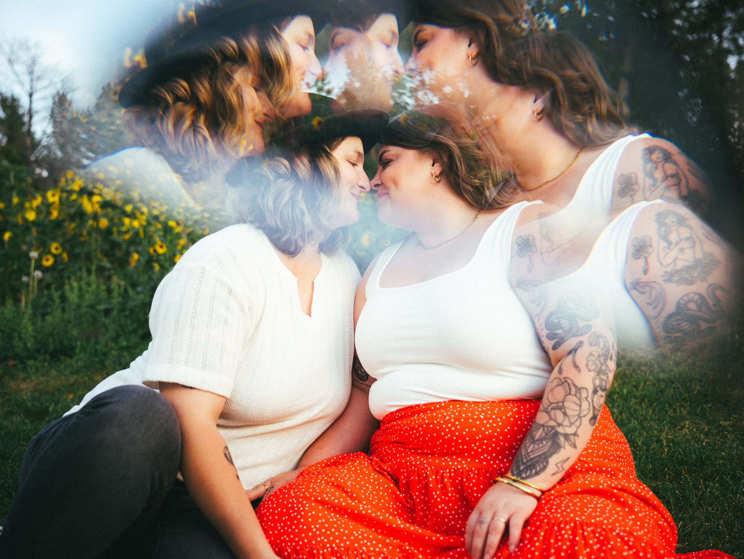 A double exposure photograph of two women with similar features, one with blonde hair and the other with dark hair and tattoos, touching foreheads and smiling at each other outdoors with yellow flowers and trees in the background.