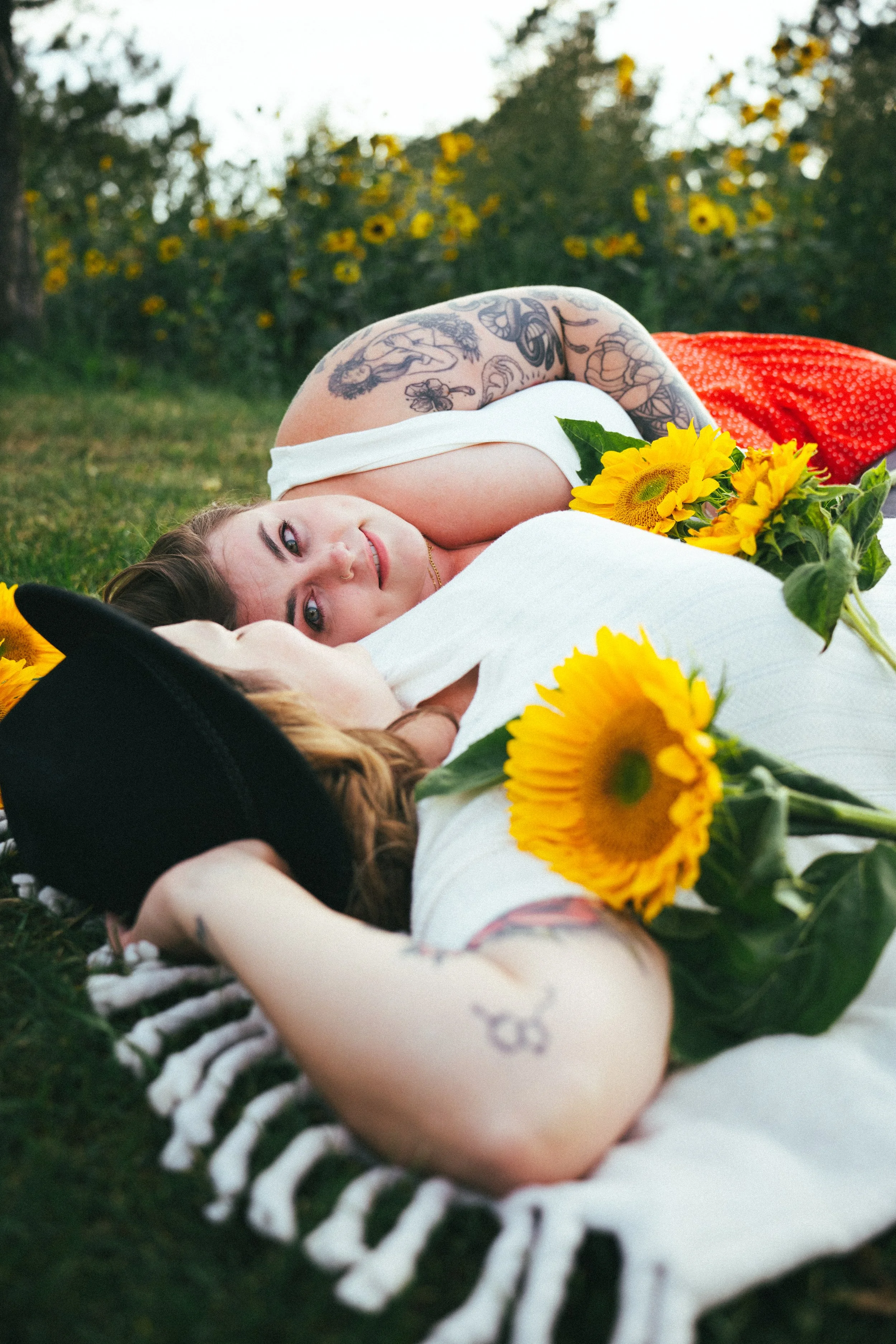 Two women lying on the grass surrounded by sunflowers, one with a black hat and tattoos, the other with short hair and a white top, gazing at each other in a park or garden setting.