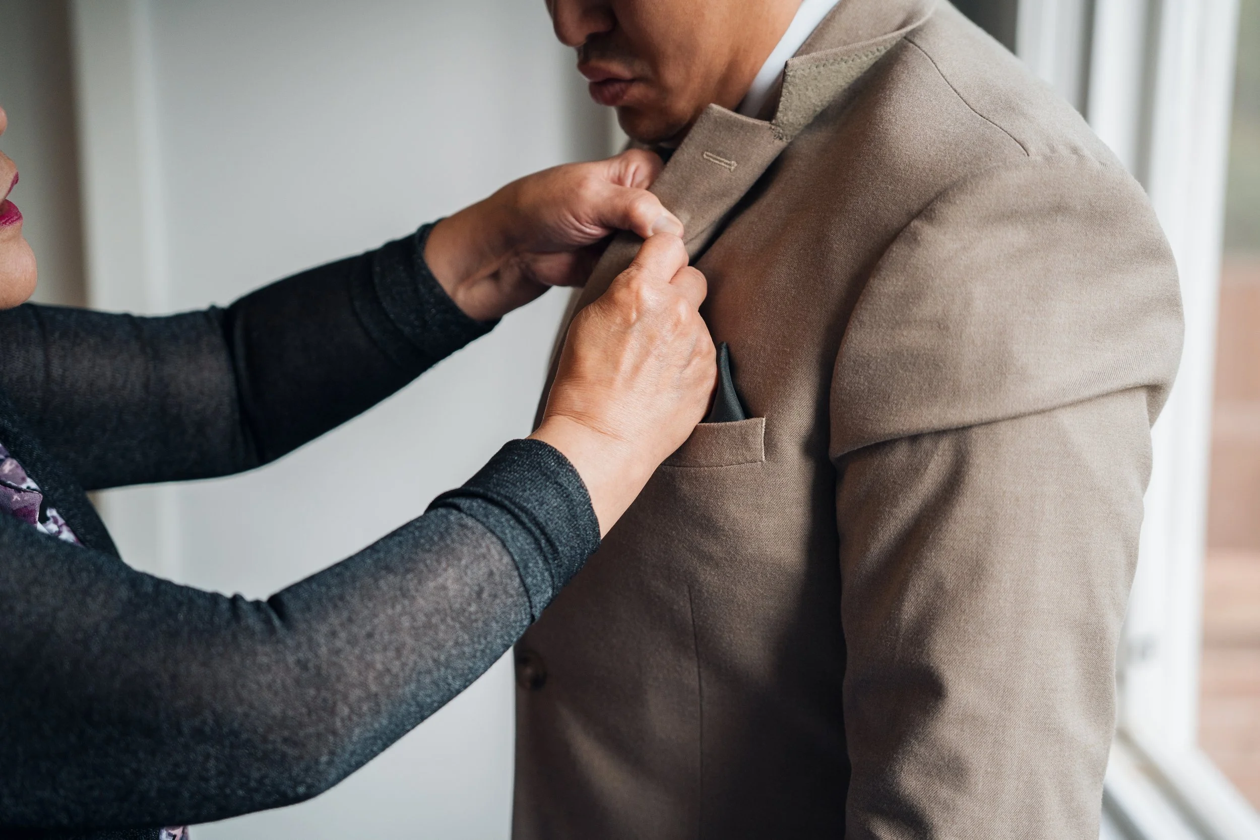Person pinning a bouton on a man's beige suit jacket near a window.