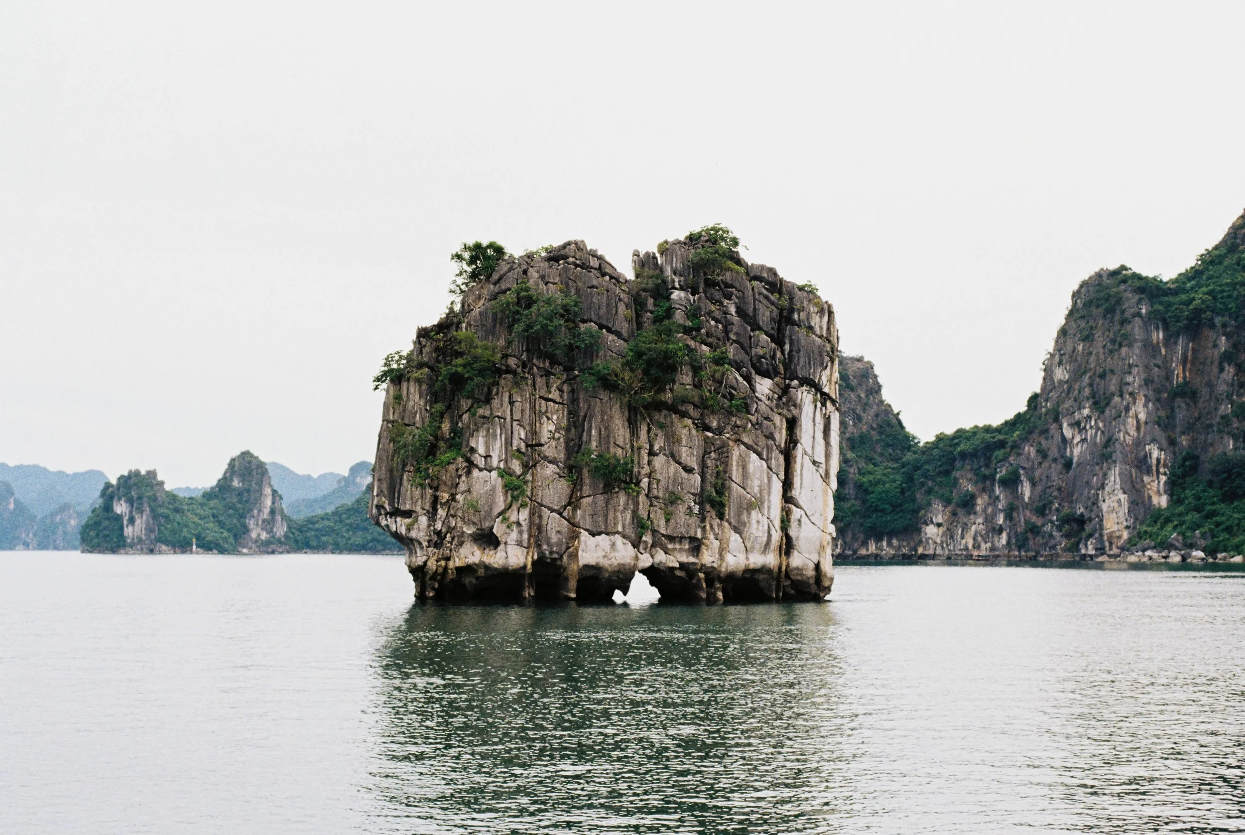 Large limestone island with greenery, surrounded by water and other islands in the background under an overcast sky.