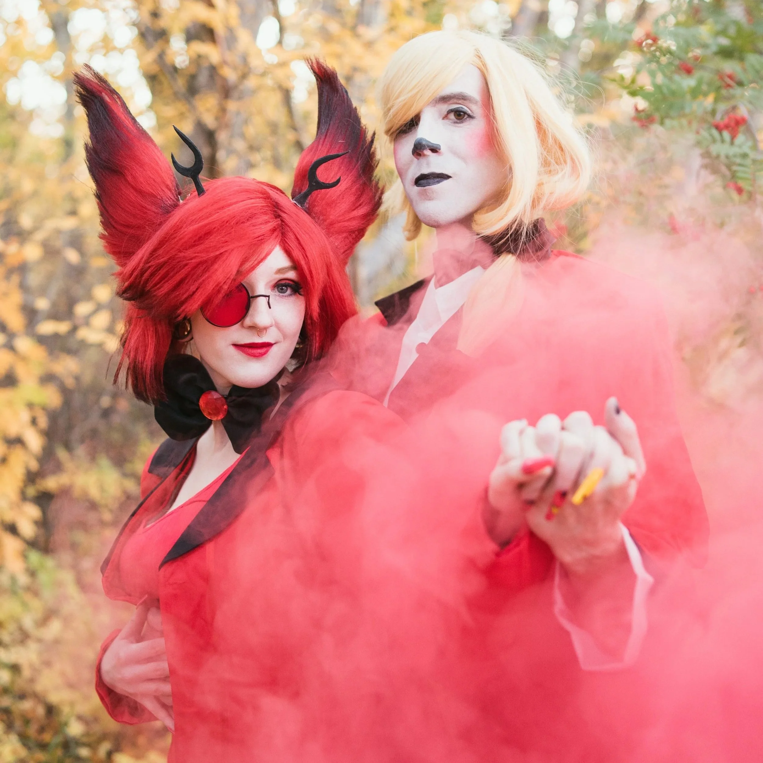 Two people in Halloween costumes, one dressed as a devil with red hair, horns, and an eye patch, and the other as a vampire with blonde hair, makeup, and a black and red cape, standing outdoors in front of autumn foliage with pink smoke in the foreground.