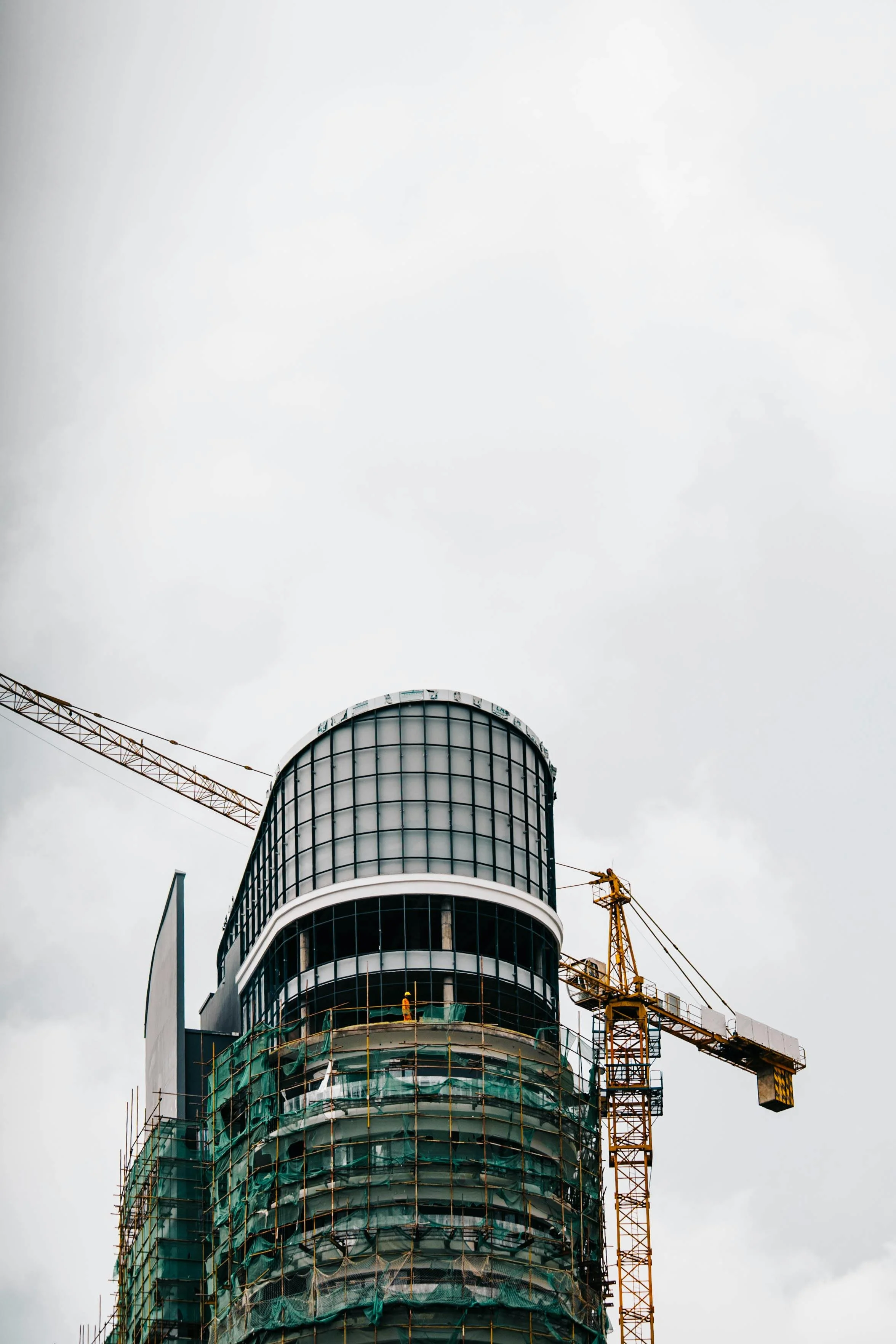 Under construction high-rise building with a glass facade and a tower crane against a cloudy sky.