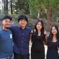 Four young adults standing outdoors in a wooded area, smiling at the camera.