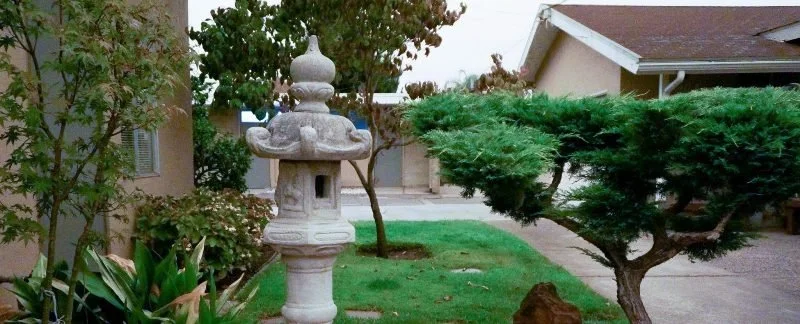 A traditional Japanese stone lantern in a suburban front yard, surrounded by bushes, trees, and a sidewalk.