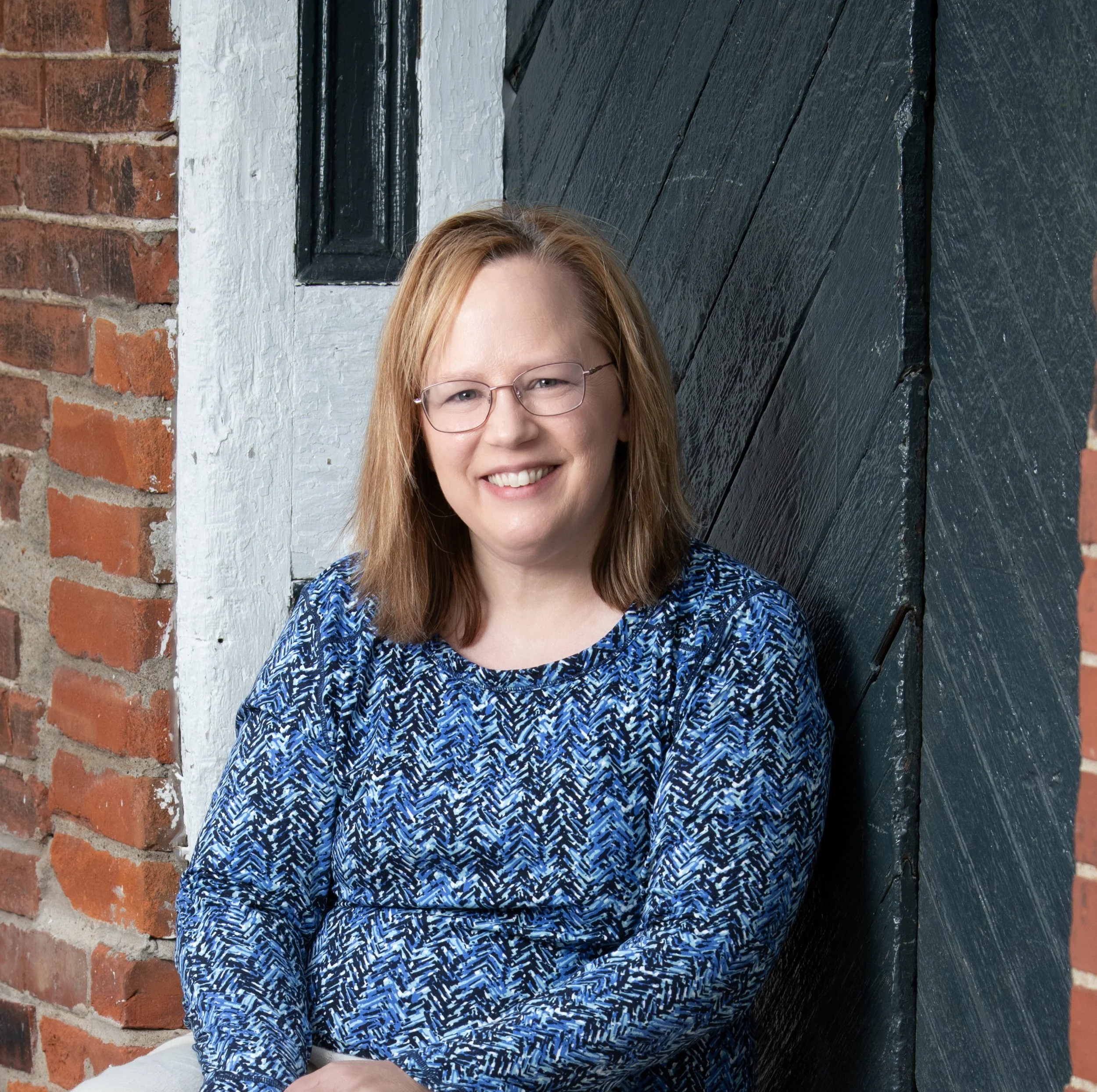 Certified Life Coach Kristin is sitting in front of a wooden door and brick background smiling warmly.