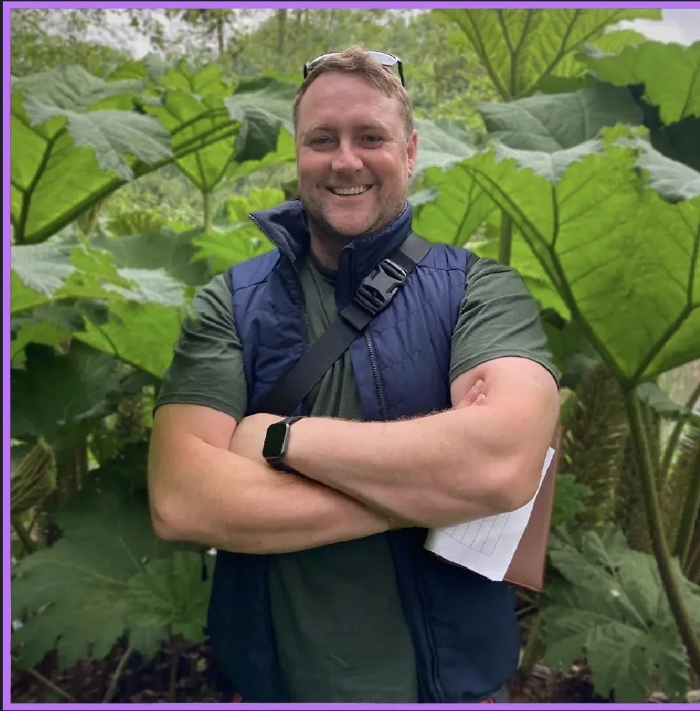 A man wearing sunglasses, a high visibility vest, red gloves, and holding equipment while standing outdoors in front of a metal fence and a sign.