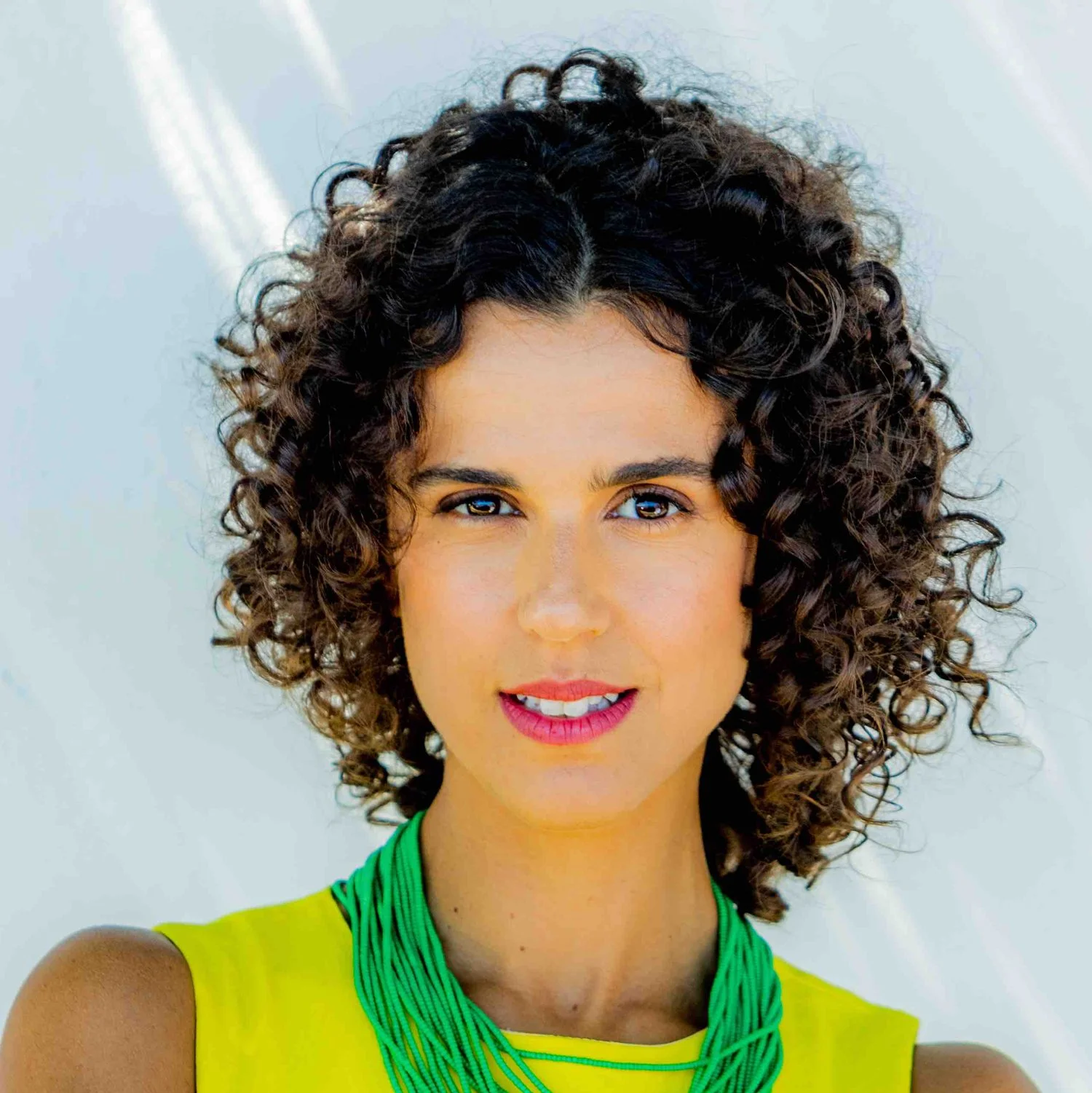 Retrato de uma mulher com cabelo cacheado, usando blusa amarela e colar verde, sorrindo para a câmera.