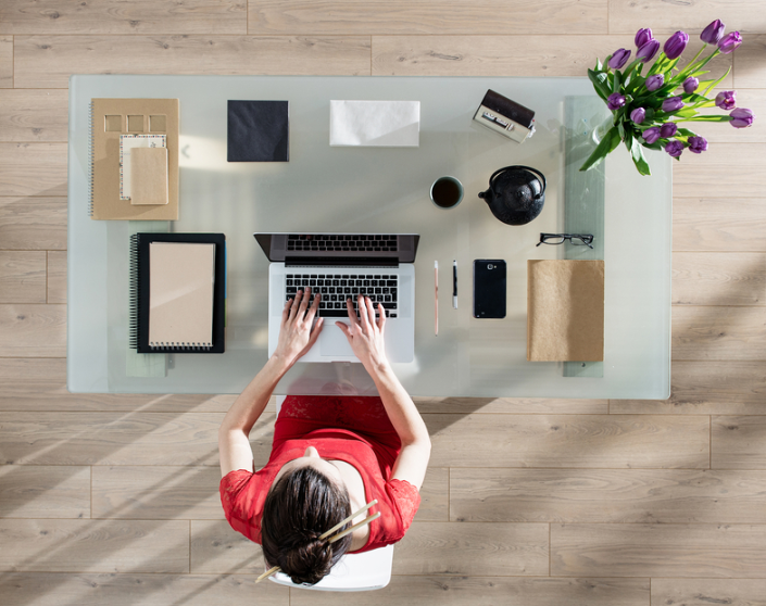 Overhead view of a woman in a red dress working on a laptop at a glass desk, with notebooks, a plant with purple flowers, a coffee mug, and other office supplies on the desk.