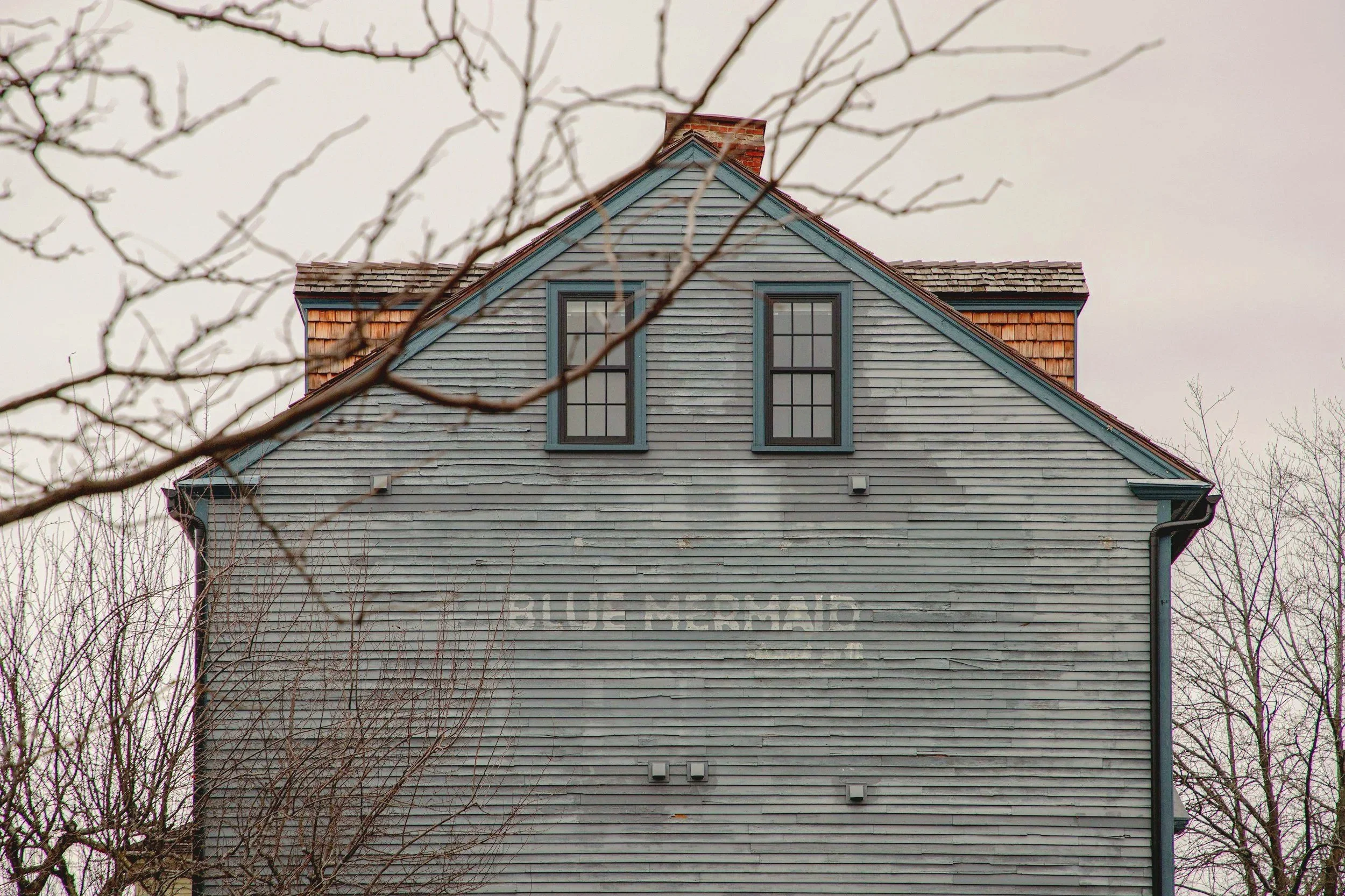 A weathered gray wooden house with two double-hung windows on the upper floor, a steep gabled roof with wood shingles, and faded painted lettering on the side that reads "BLUE MERMAID". Bare tree branches are in the foreground, and the sky is overcast.