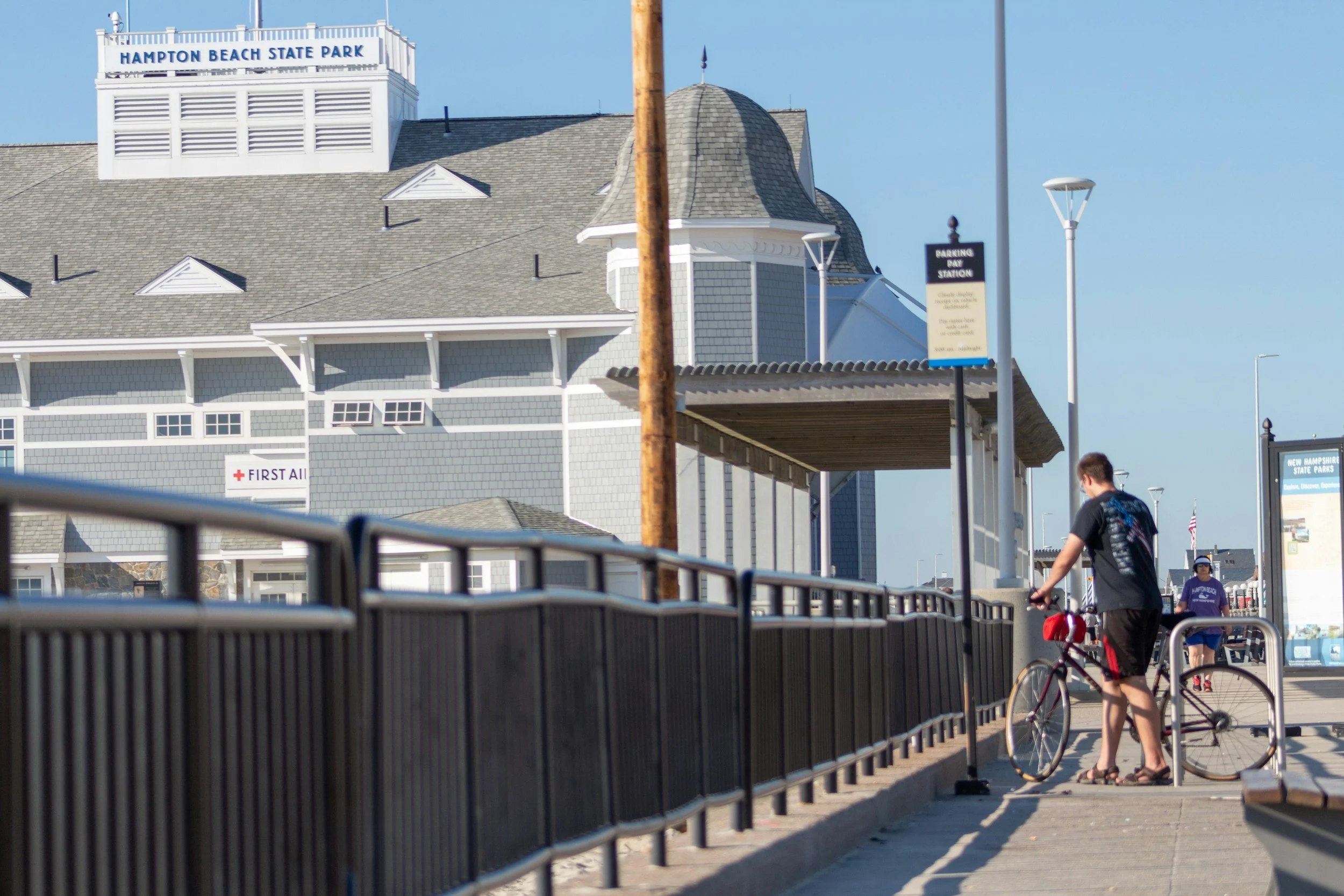 People walking and cycling along a sidewalk outside Hampton Beach State Park with a beach building and shelter in the background on a sunny day.