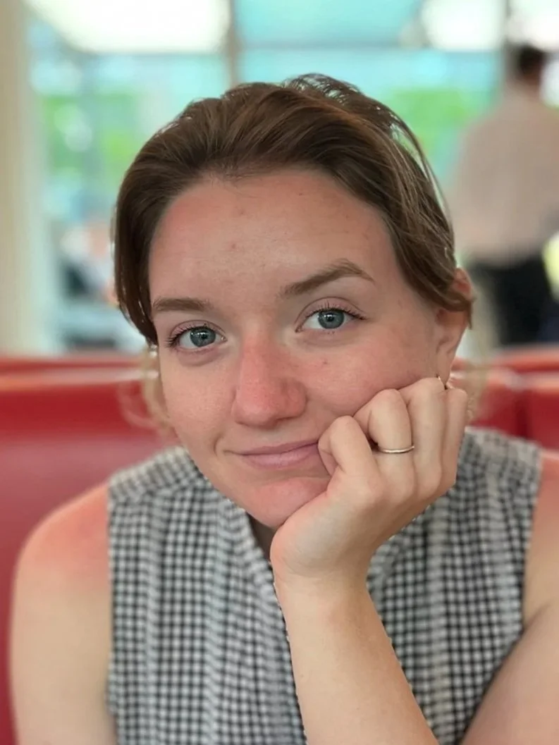 A young woman with short brown hair and blue eyes resting her face on her hand, sitting in a diner or cafe with red booths and a blurred background.