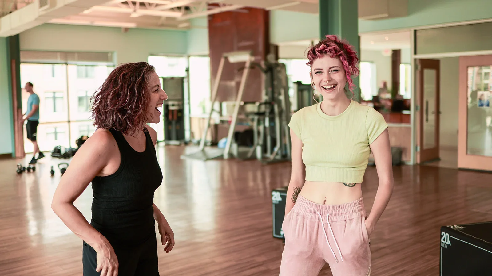 Two women laughing and talking in a gym. One woman has curly red hair, is wearing a black tank top, and the other has short pink curly hair, is wearing a yellow crop top and pink sweatpants. A man is in the background near gym equipment.