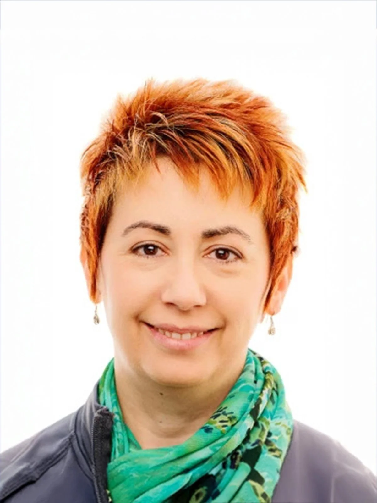 Portrait of a woman with short, curly, orange hair smiling, wearing a black jacket and a green patterned scarf, against a white background.