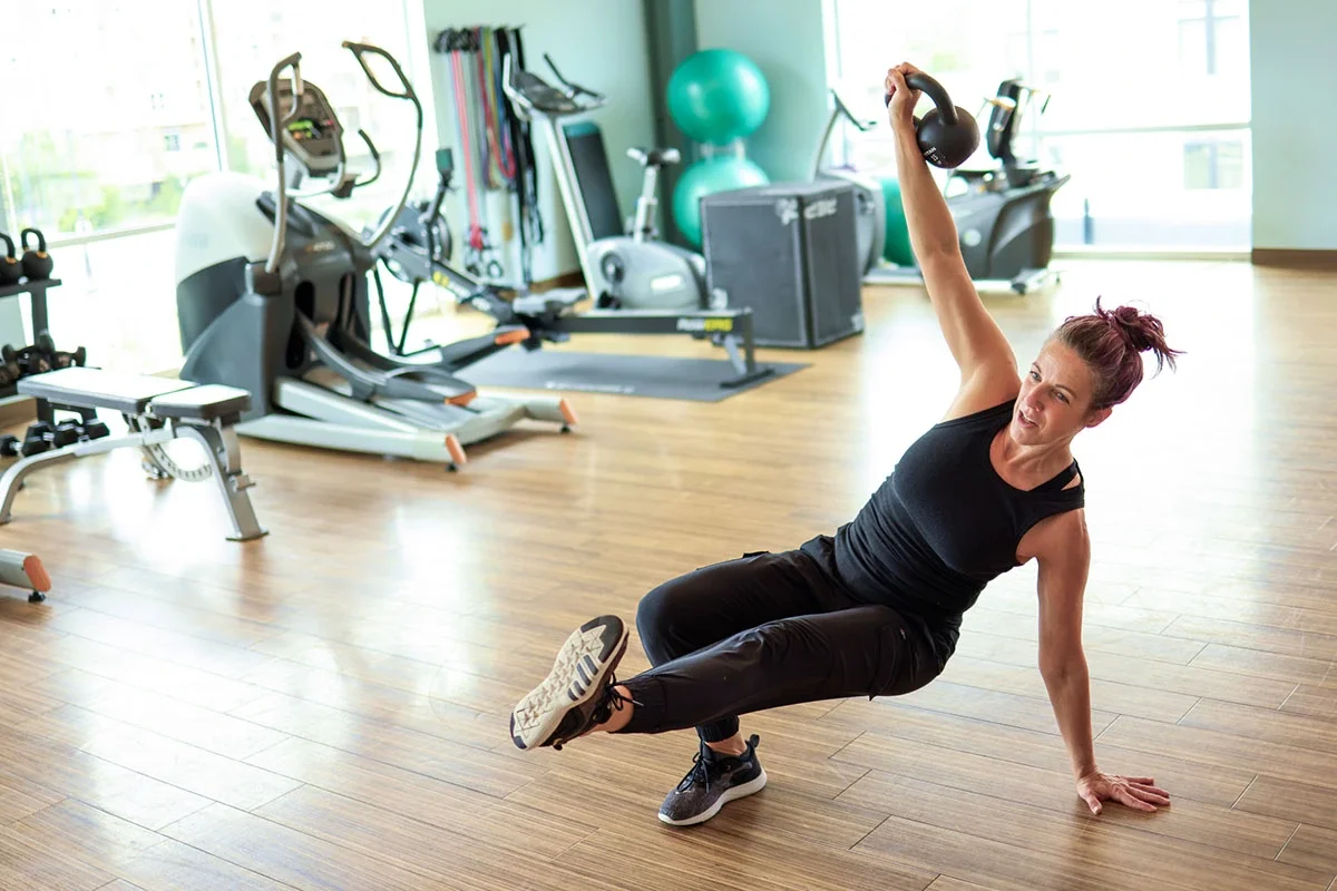 A woman doing a side plank exercise with a kettlebell in a gym with exercise equipment, including a rowing machine and stability balls.