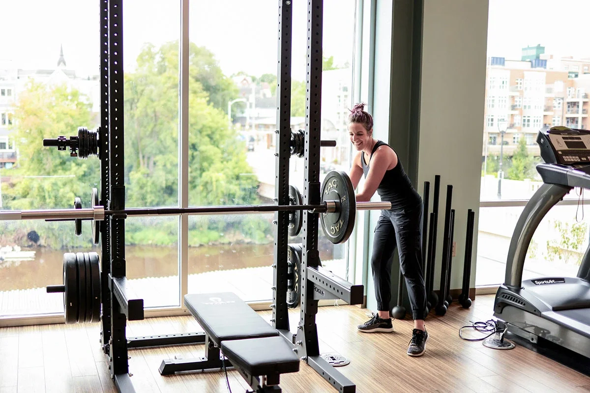 A woman in black workout clothes lifting a barbell with weights in a gym near large windows showing a city view.