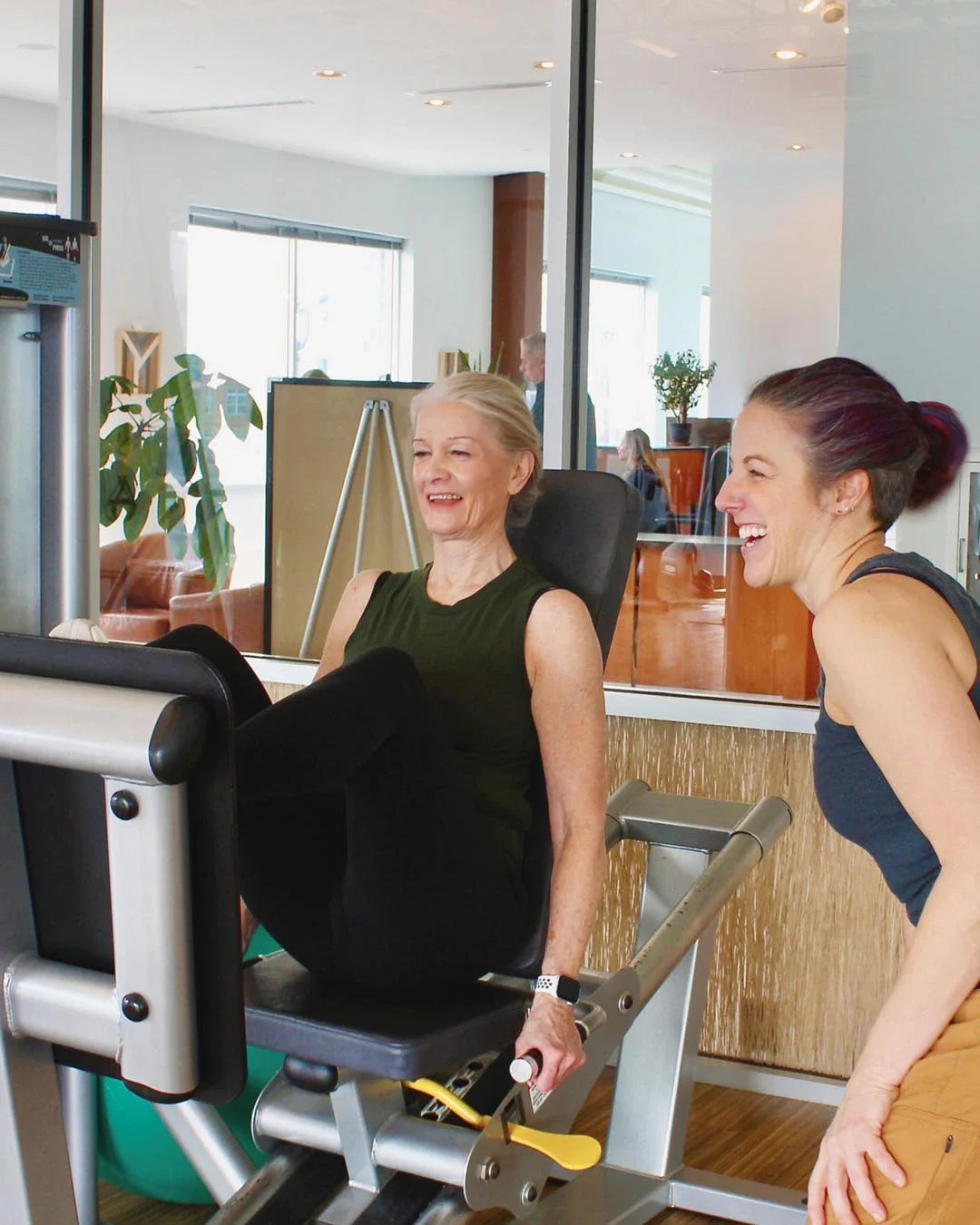 A woman in her 60s or 70s with short blonde hair sitting on an exercise machine, smiling, with a younger woman laughing beside her in a fitness center.