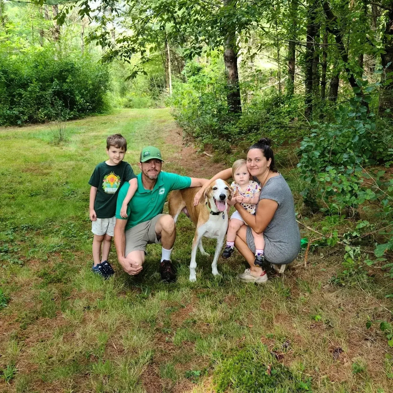 Family of four with their dog in a lush green forest, squatting on grass and looking at the camera.