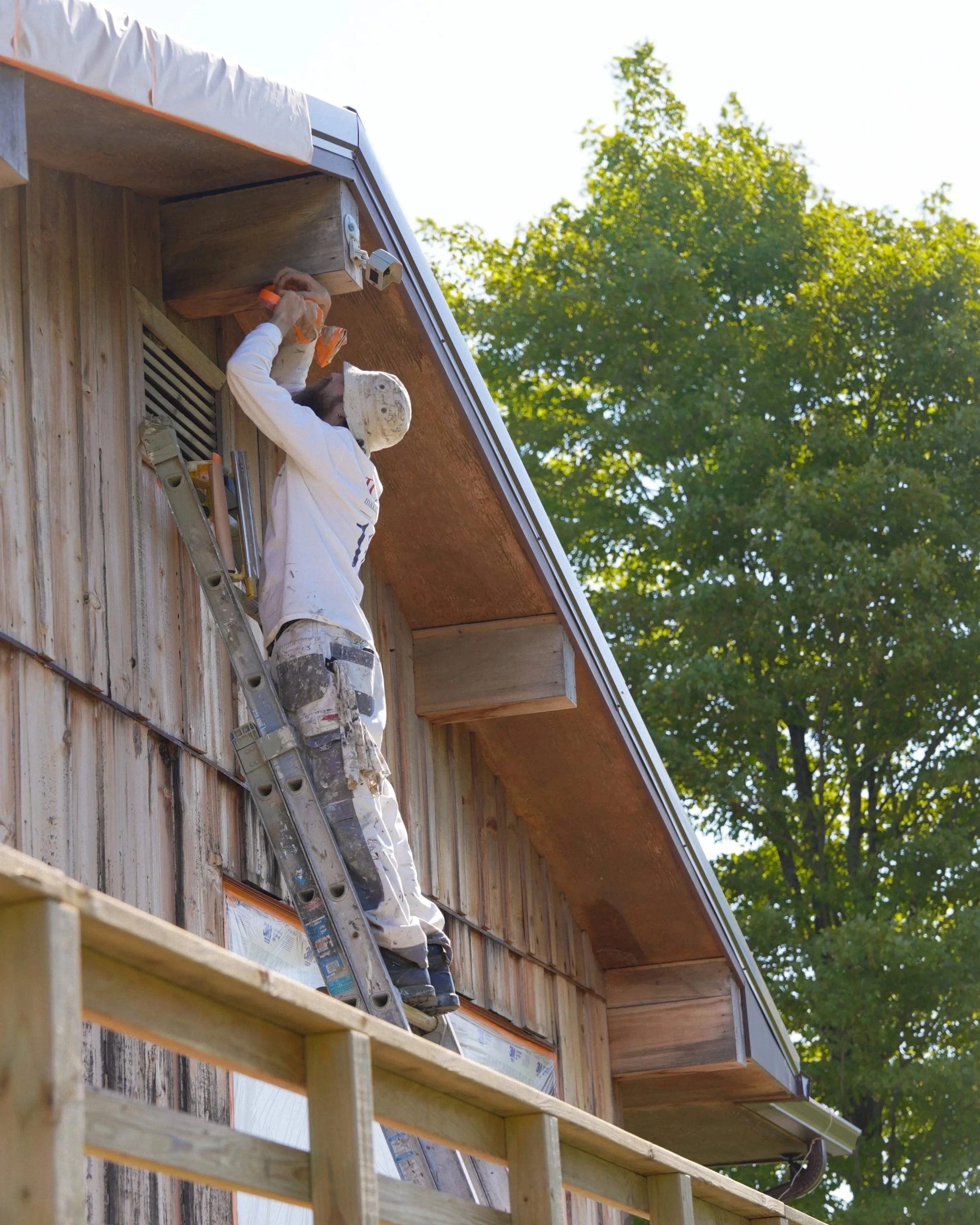 A person wearing a white hoodie and cap is working on the exterior of a wooden house, standing on a ladder and using a tool on the eaves under the roof, with trees visible in the background.