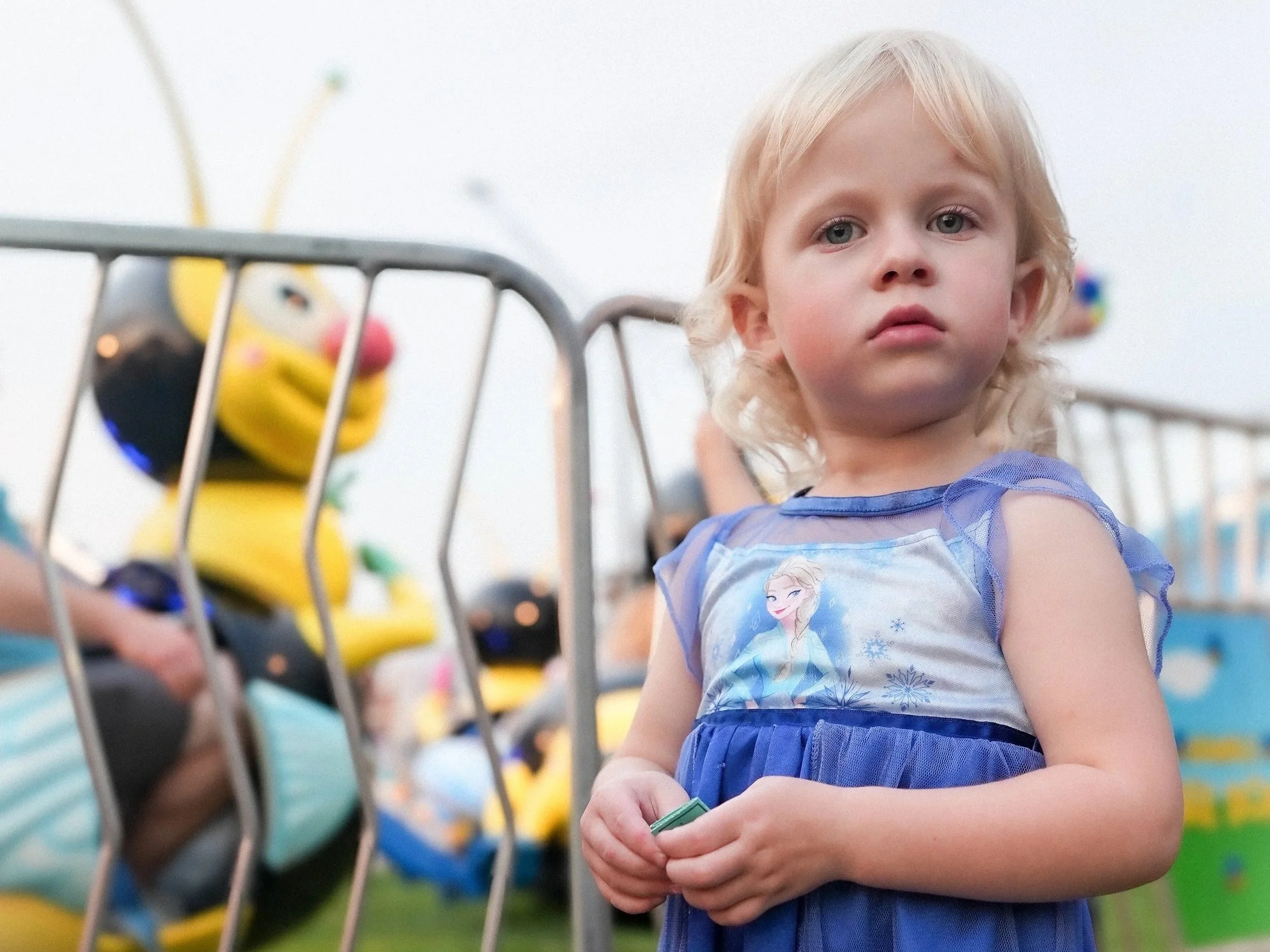 A young girl with blonde curly hair and blue eyes standing in a colorful indoor play area, wearing a blue dress with an Elsa from Frozen design, holding a green toy.