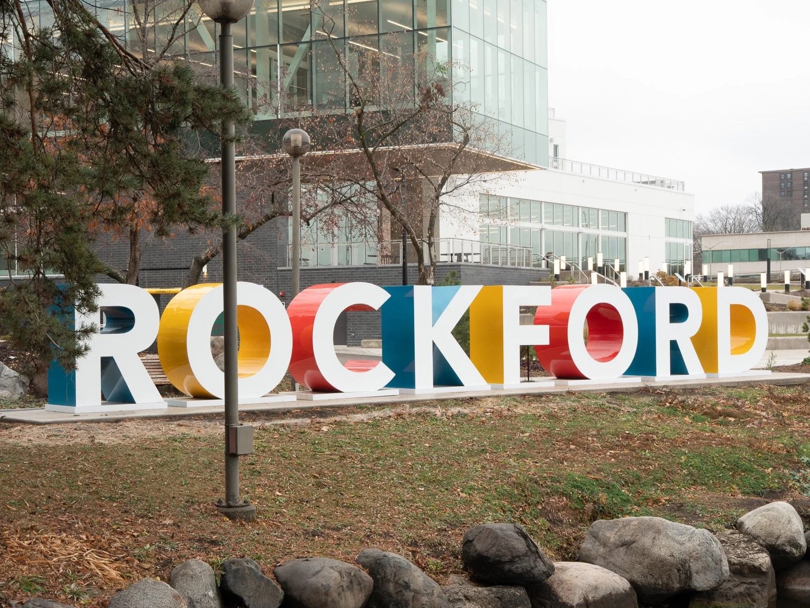 Colorful 3D sign spelling 'ROCKFORD' on a grassy area with rocks in the foreground, trees with leaves and a modern glass building in the background.