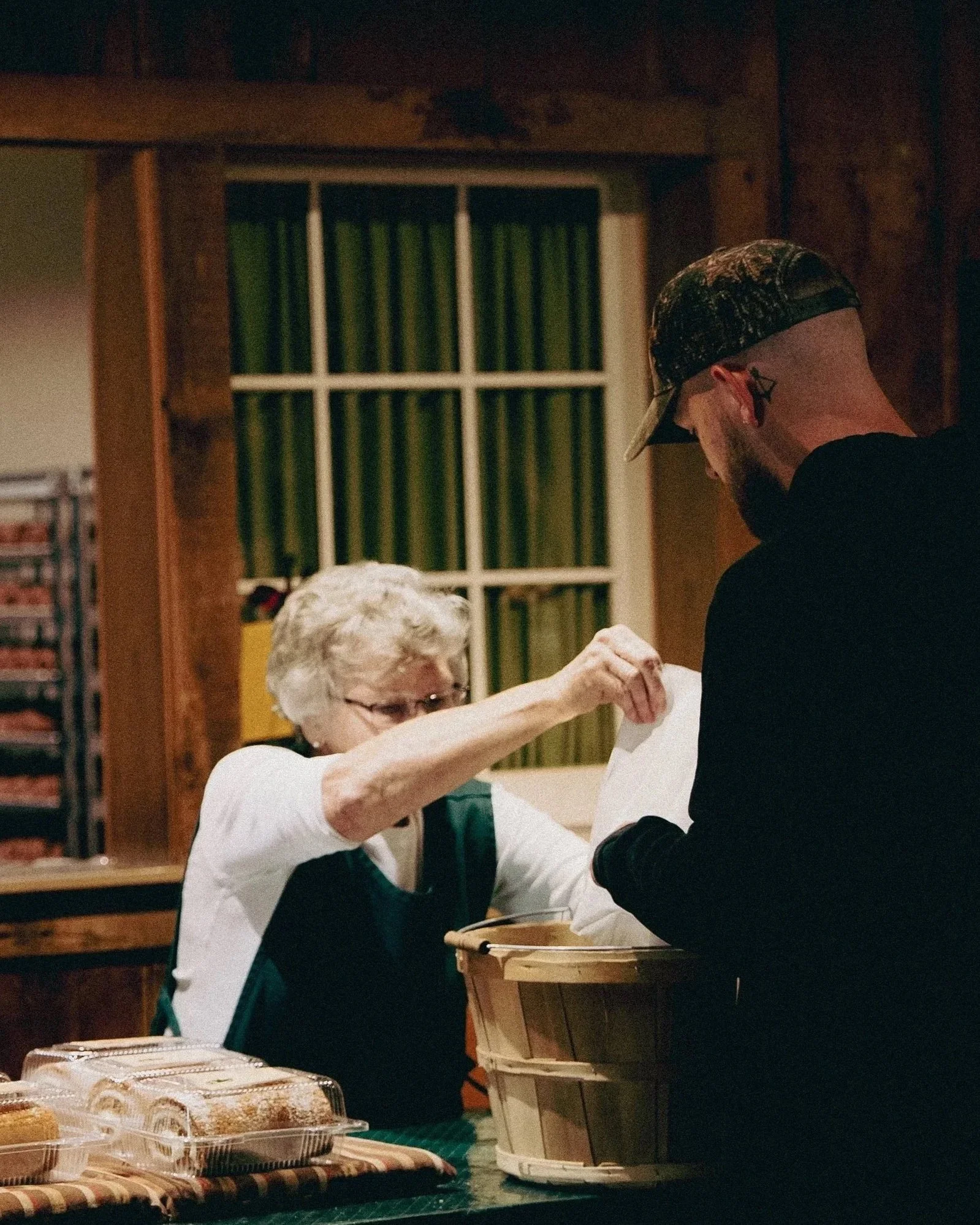 An elderly woman with glasses and short white hair wearing a green apron and a white shirt with rolled-up sleeves is reaching out to a man with a beard, wearing a cap and a black shirt, in a cozy indoor setting, possibly a kitchen or a bakery. The table in front of them has food packages and baked goods.