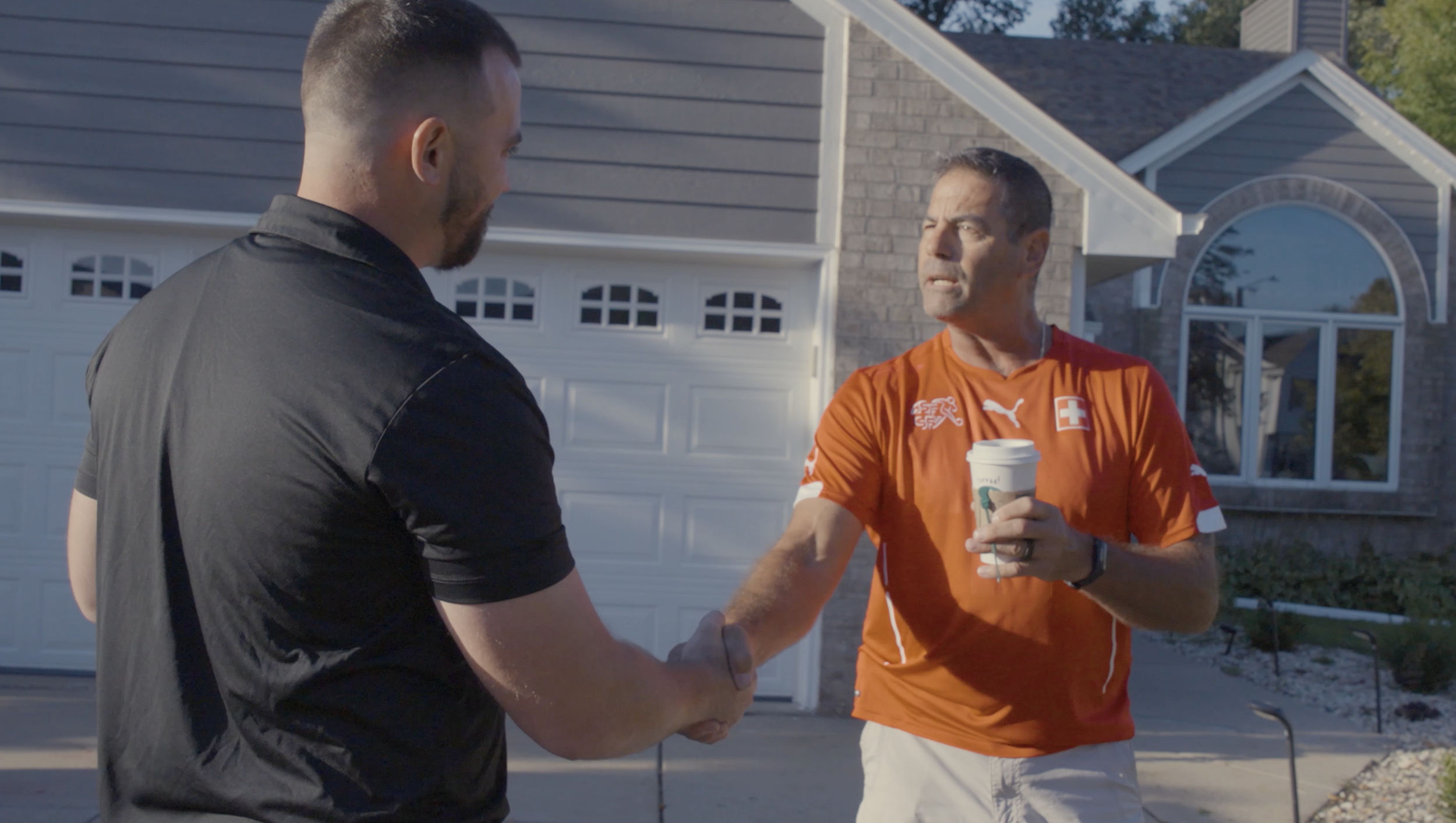Two men shaking hands outdoors in front of a house, one wearing a black shirt and the other in an orange sports shirt holding a coffee cup, during daytime.