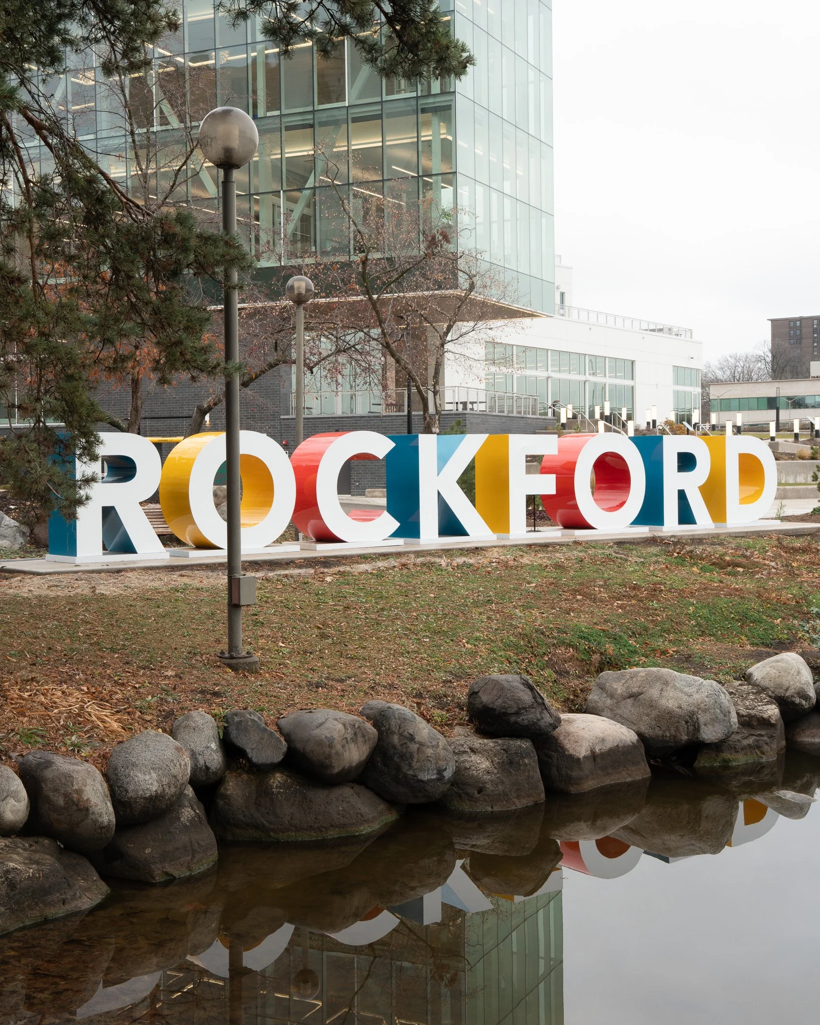 Large colorful rocks spelling out 'ROCKETOP' are placed near a calm body of water, with a modern glass building, trees, and street lamps visible in the background.