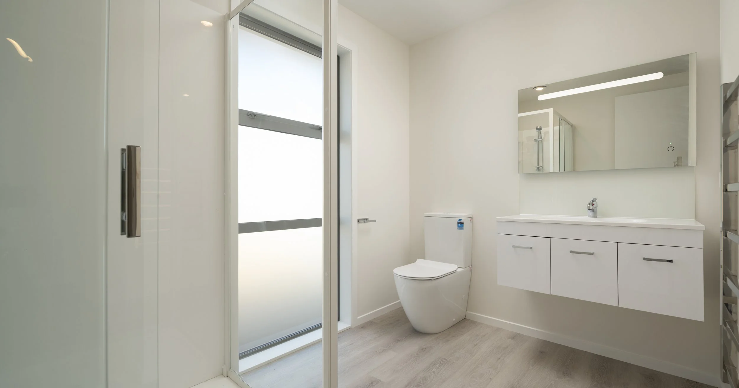 Modern minimalist bathroom with white walls, a large mirror above a white vanity with a faucet, a toilet, and a frosted glass door leading outside in Blenheim New Zealand