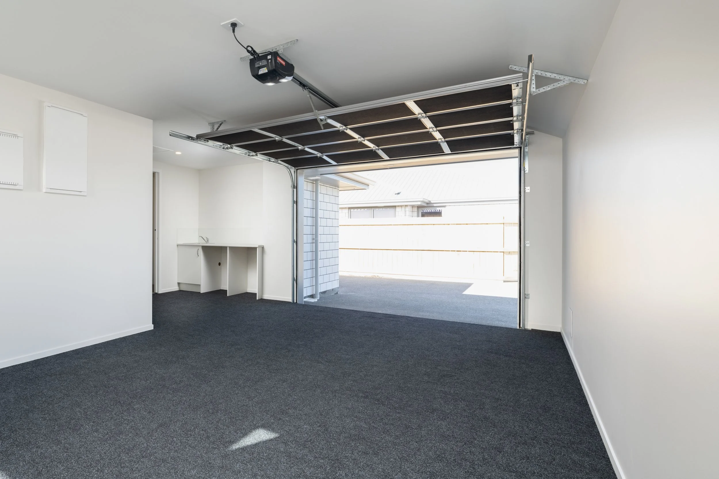An empty garage with a partially open garage door, gray carpet flooring, white walls, and a small built-in countertop with a sink. There is a ceiling-mounted garage door opener in Blenheim New Zealand