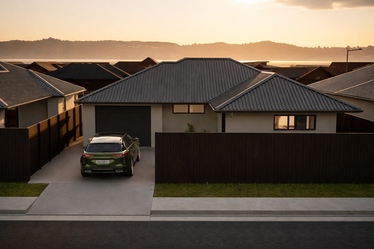 Modern house with gray metal roof, parked black SUV in driveway, surrounded by brown wooden fence, neighboring houses, and distant mountains under a sunset sky.