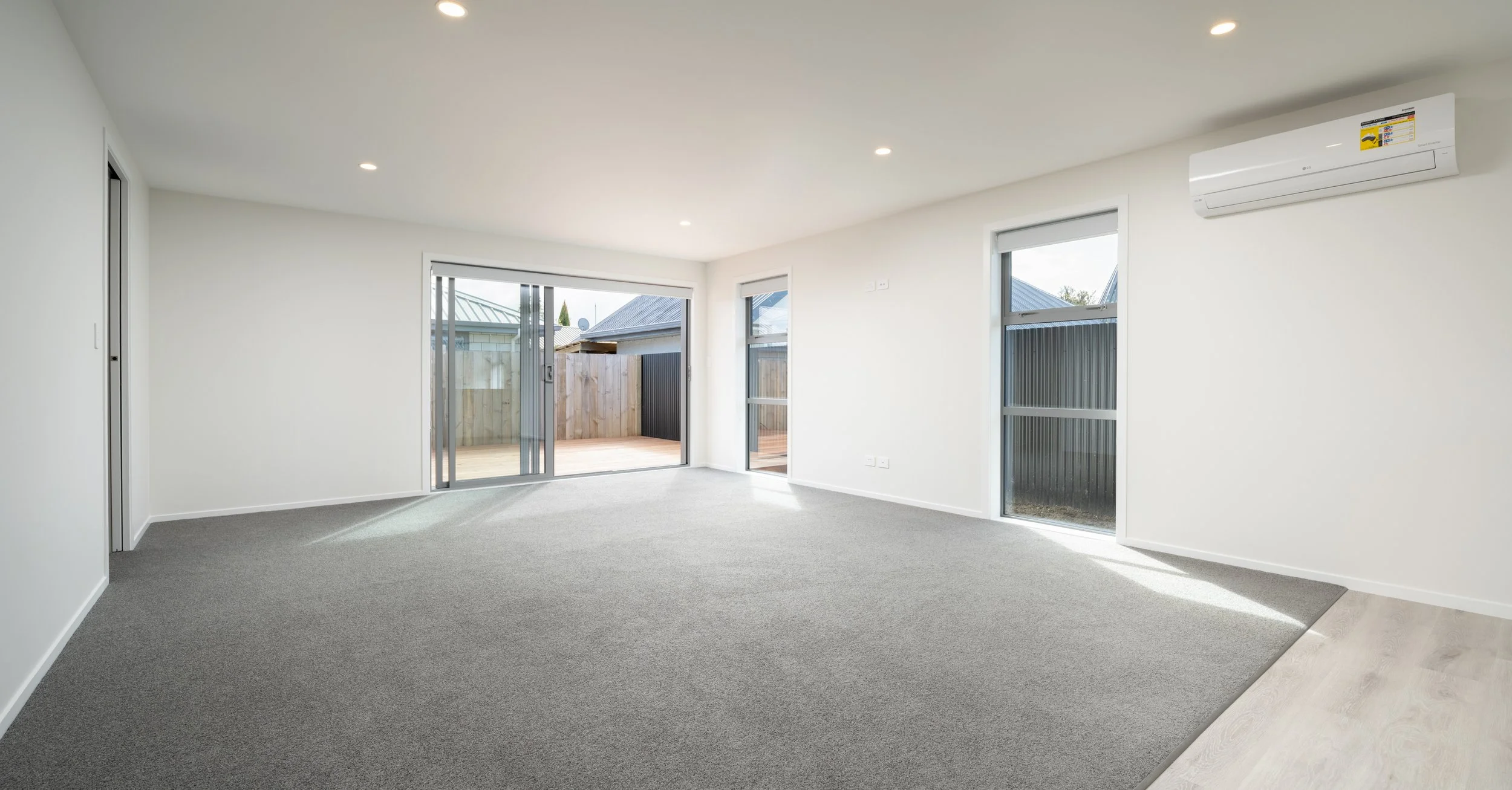 Empty living room with gray carpet, white walls, large windows, sliding glass door leading to outdoor patio, and a wall-mounted air conditioner  in Blenheim New Zealand
