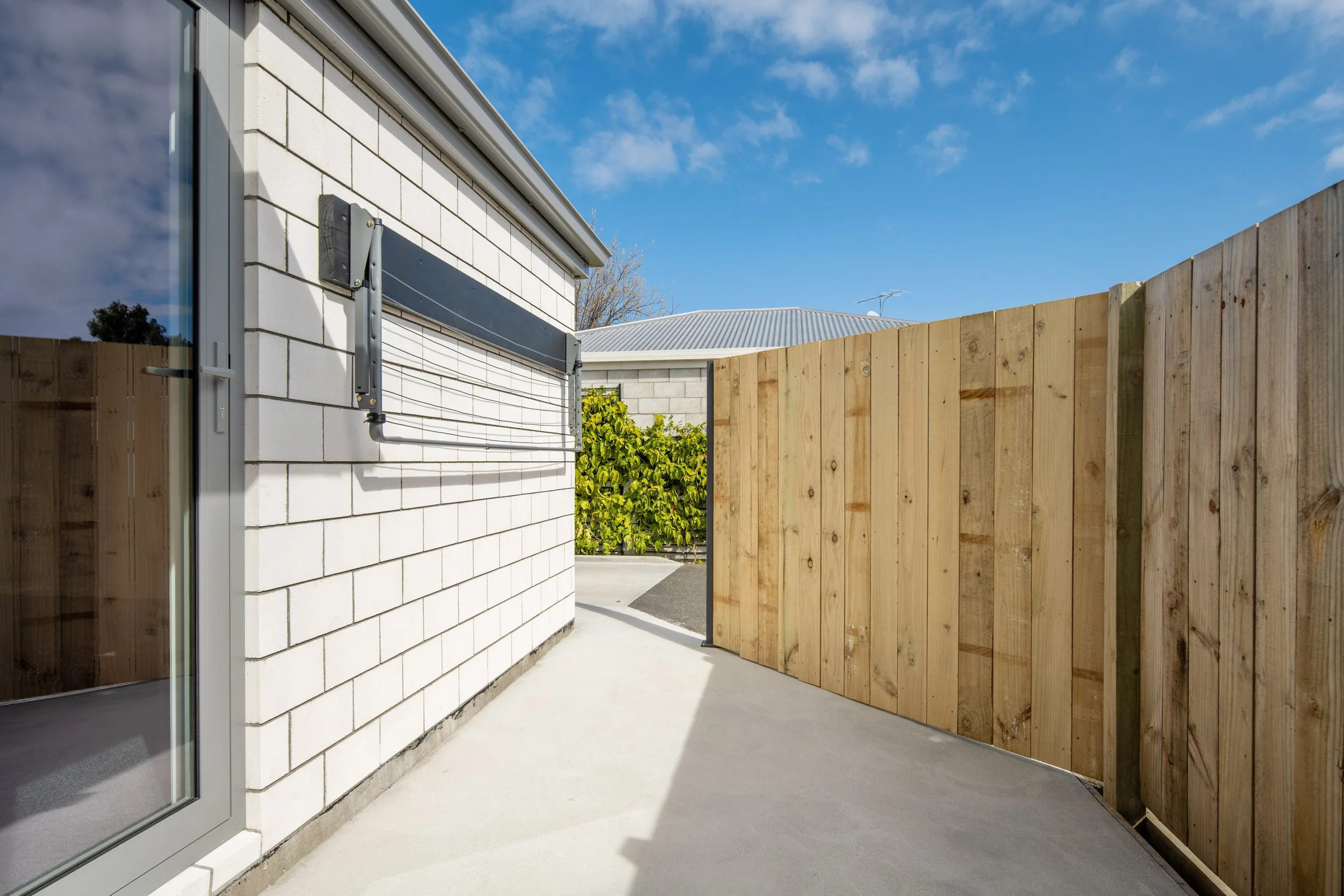 A backyard with a white brick house, a wooden privacy fence, and a concrete patio under a partly cloudy blue sky  in Blenheim New Zealand