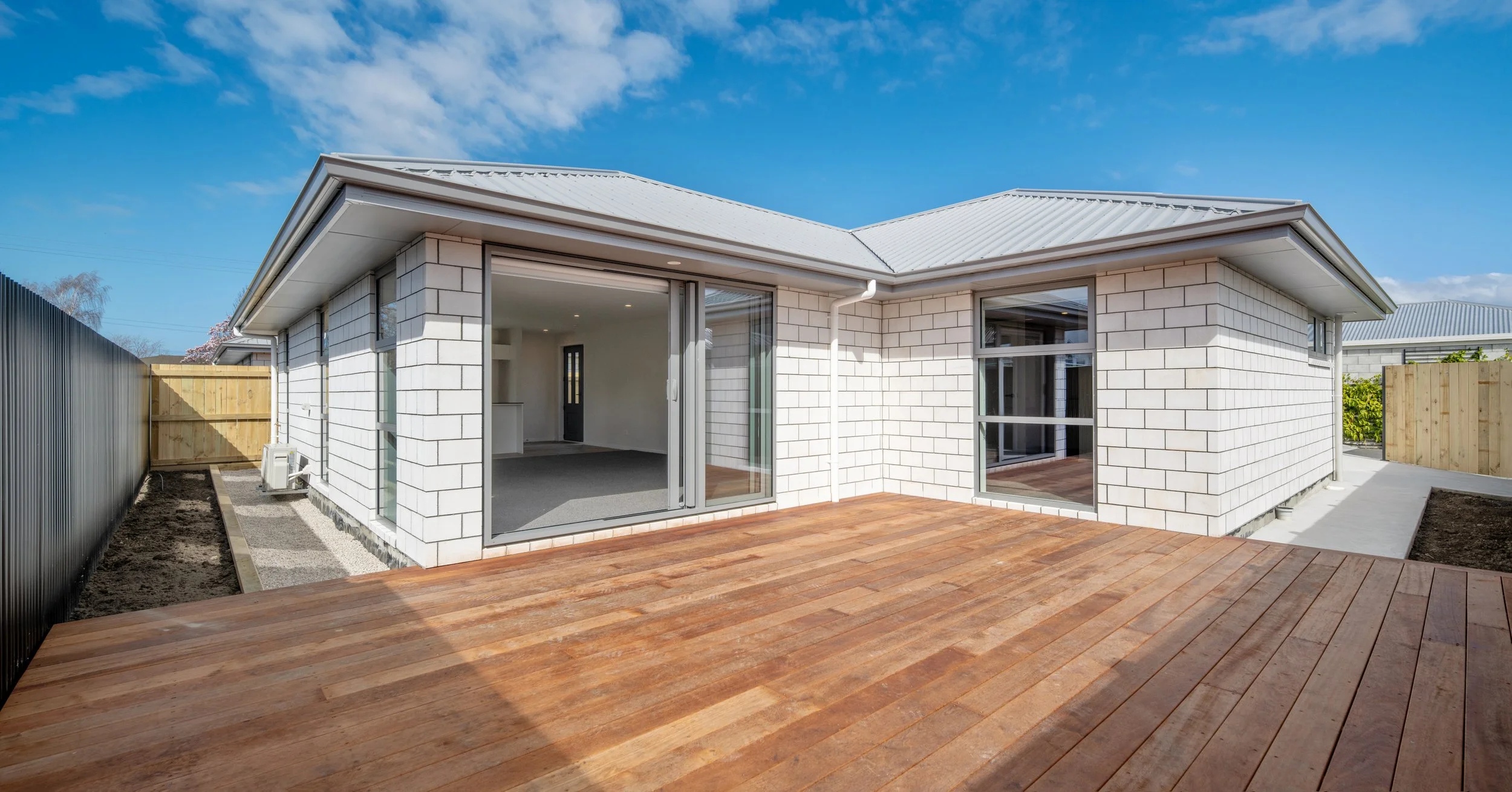 Modern house with white brick exterior, large glass sliding door, and wooden deck, under a blue sky with some clouds in Blenheim New Zealand