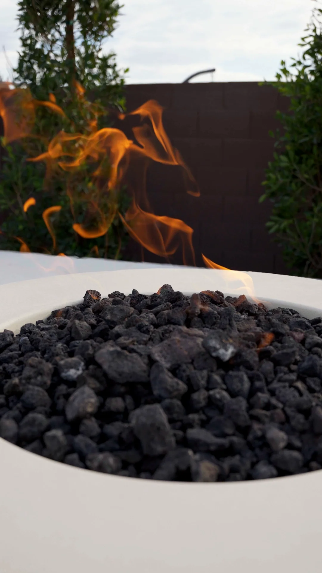 Close-up of a fire pit filled with black rocks, with flames rising up behind green bushes and a dark wall in the background in Gilbert, AZ
